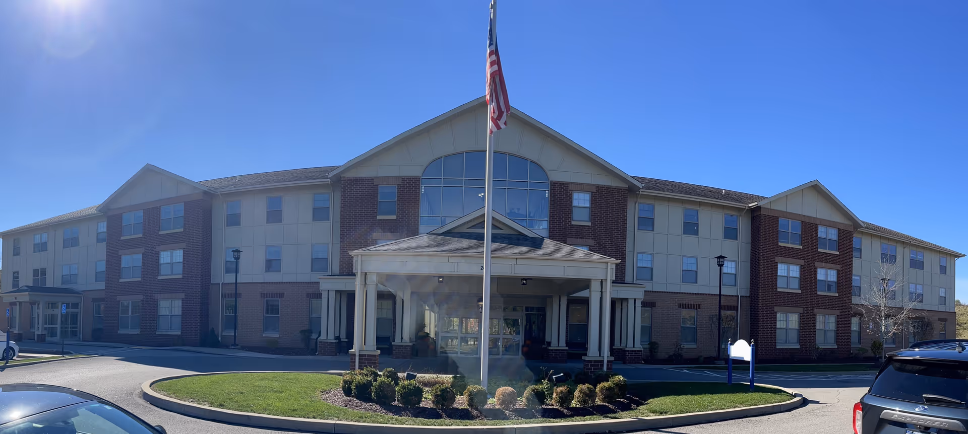 Front exterior view of a three-story senior living facility building with a covered entrance, an American flag on a flagpole in front, and a clear blue sky.