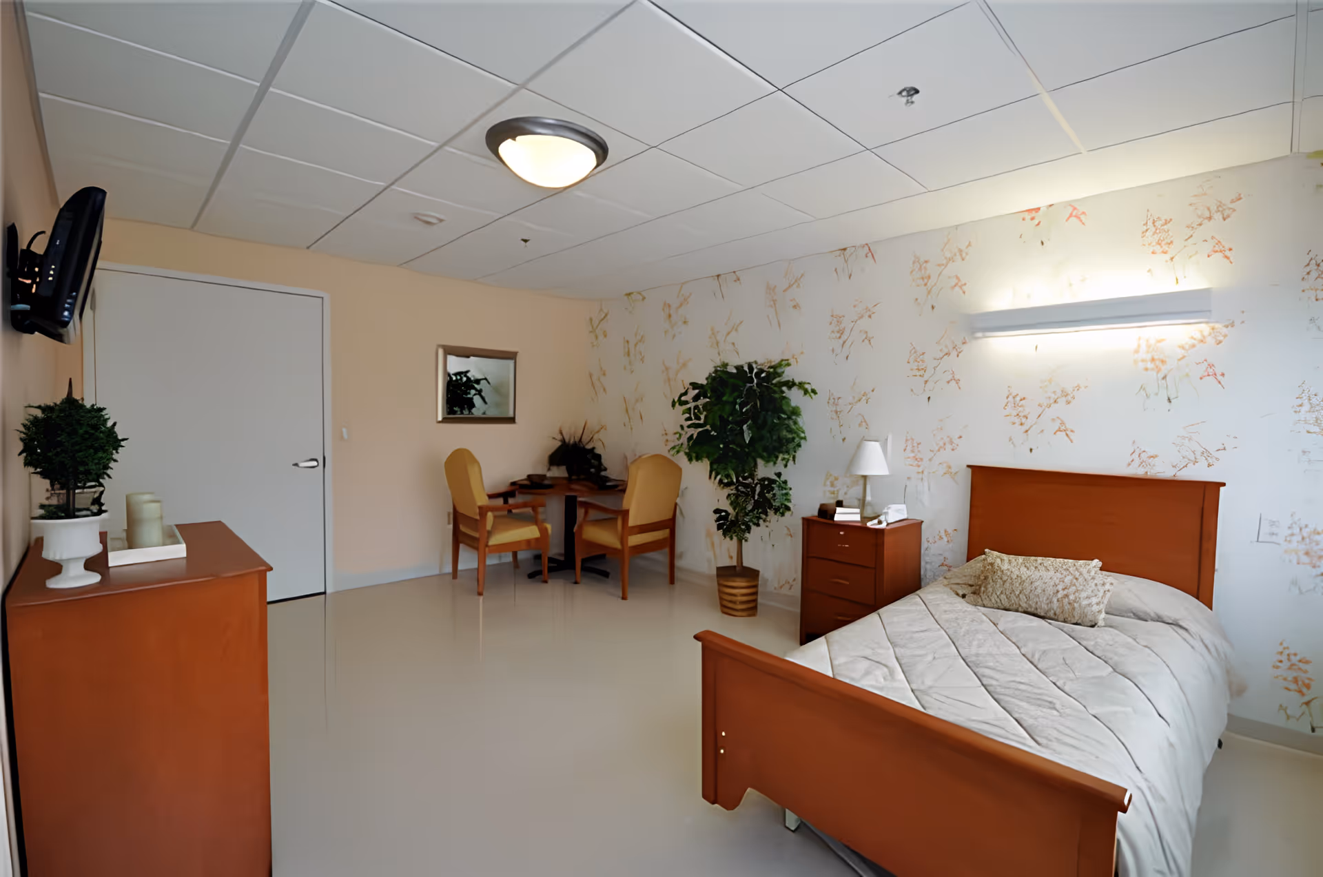 A bright private bedroom in a senior facility with a single wooden bed, nightstand, small table with chairs, dresser, and potted plants.