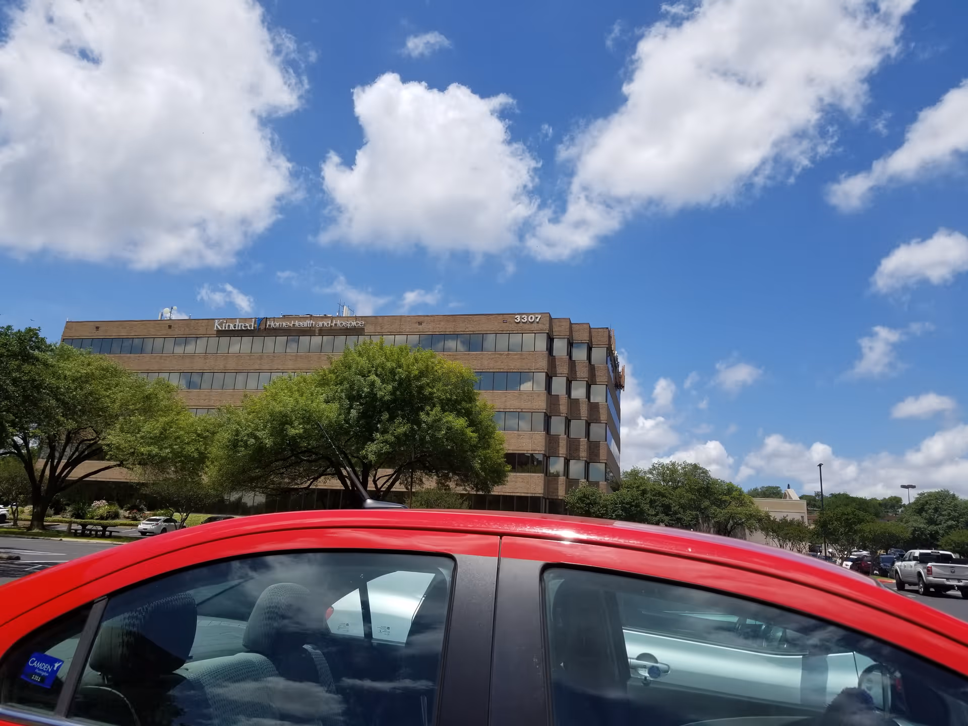 Exterior view of a brick senior care facility with trees, parked cars, and blue sky.