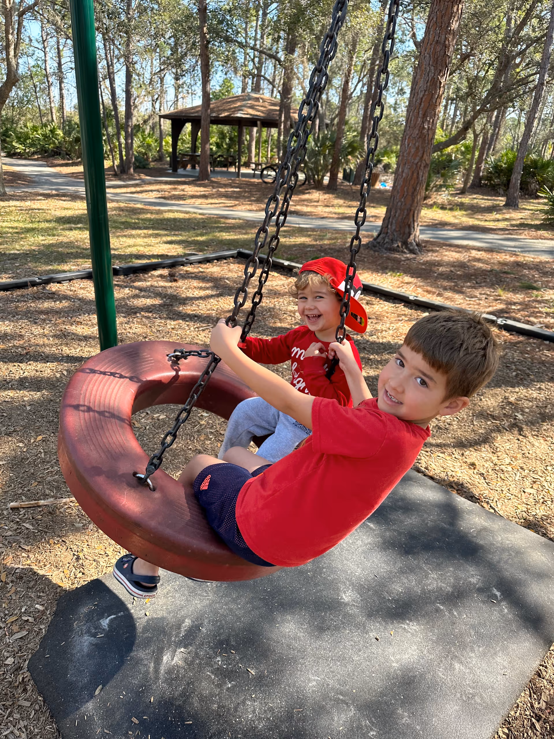 Two children sit on a red circular swing in a shaded outdoor park.