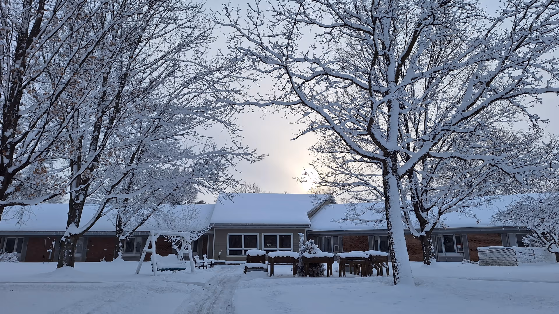 Snow-covered exterior of a single-story senior living facility with leafless trees and outdoor seating.