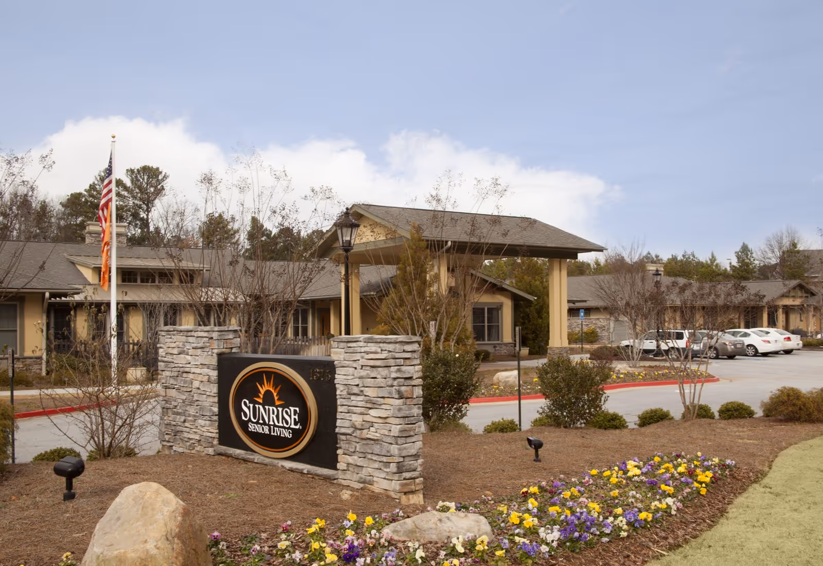 Sunrise Senior Living exterior with sign, flagpole, flowers, and parked cars at the entrance