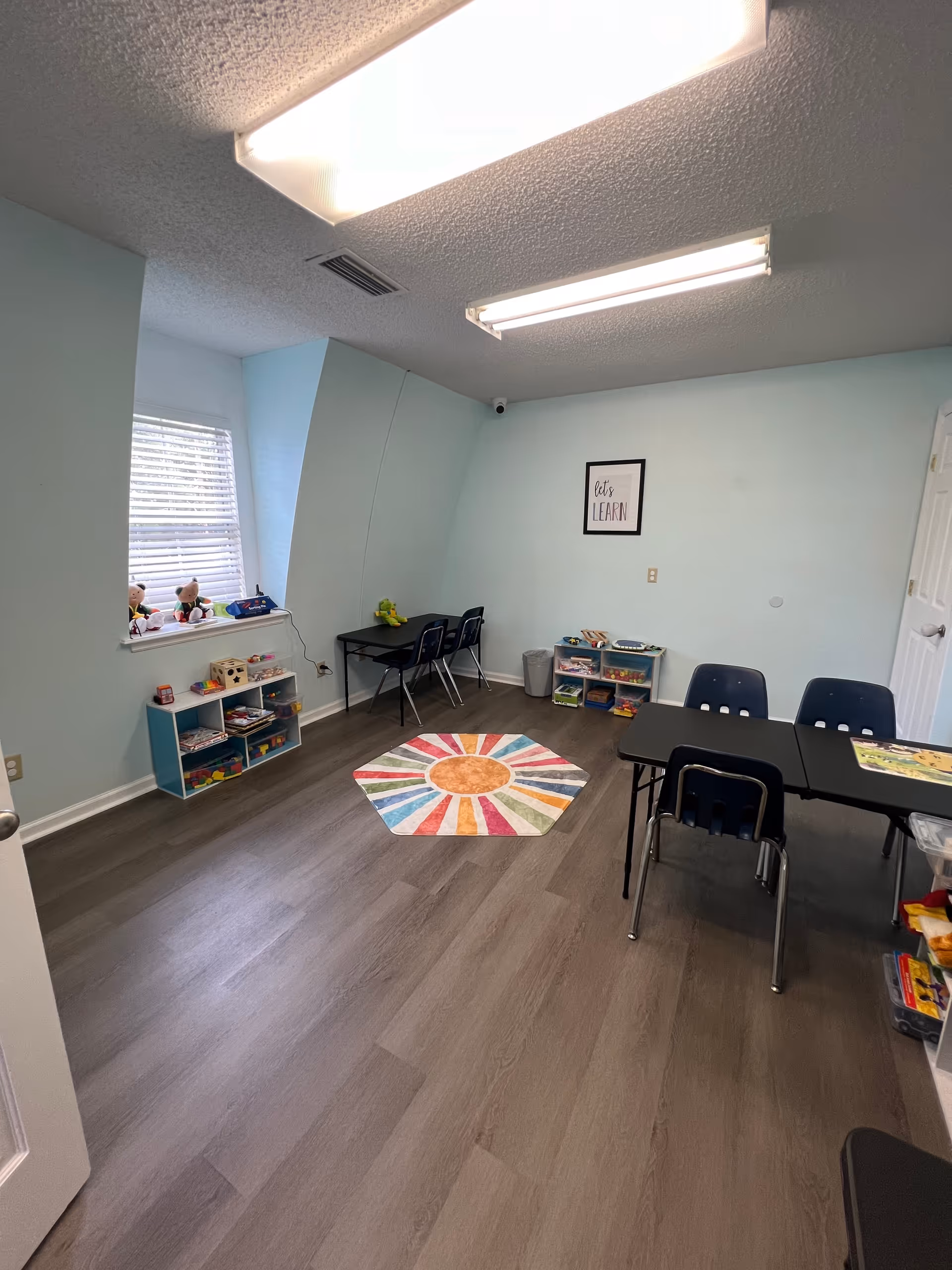 Interior playroom with pastel walls, colorful rug, toy shelves, and small tables.