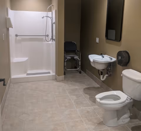Accessible bathroom with shower, sink, toilet, and a wheelchair in the corner.