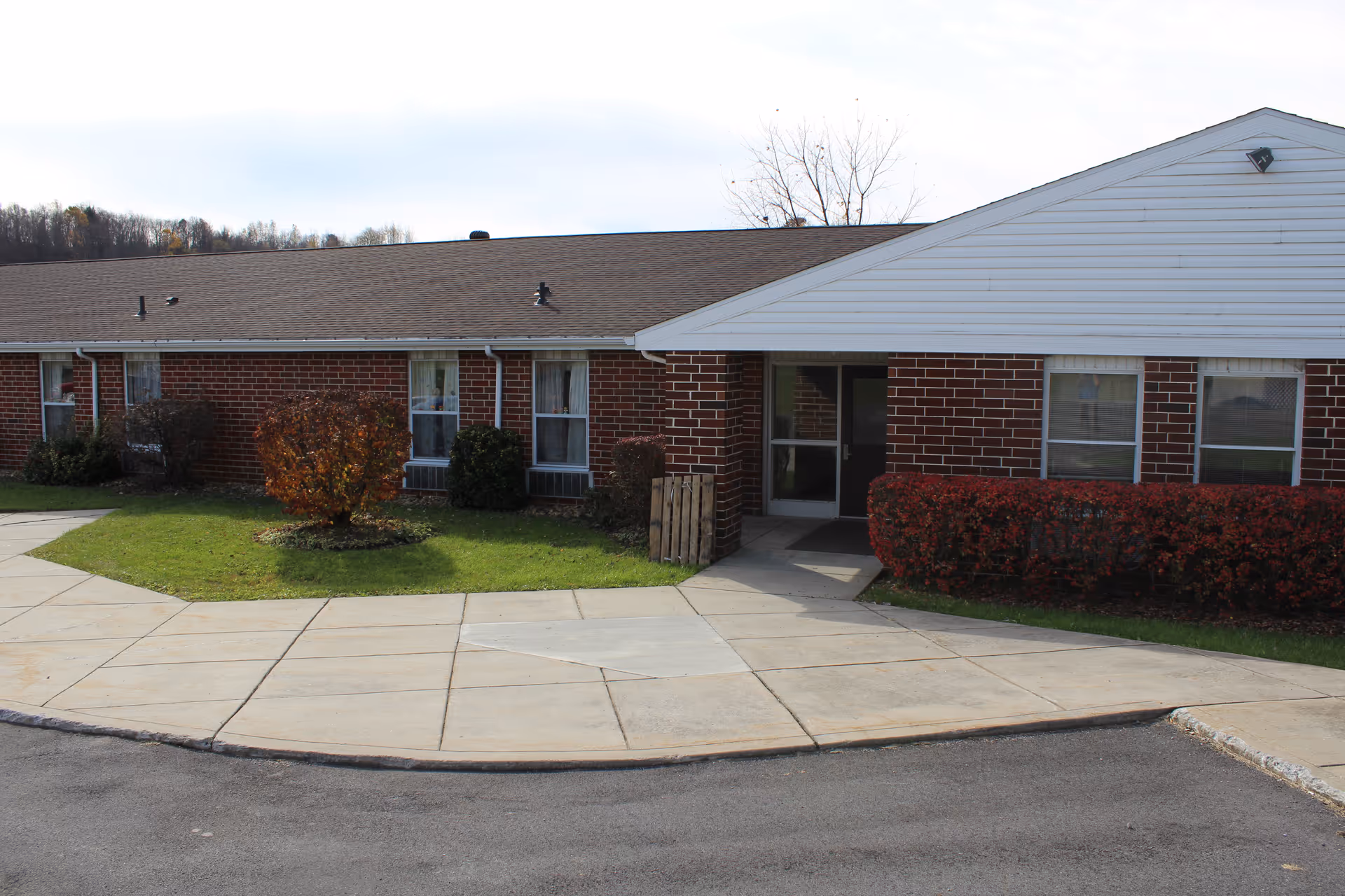 Exterior view of a single-story brick building with a sloped roof, several windows, and a main entrance door. The building is surrounded by a concrete sidewalk, green grass, and trimmed bushes with some trees in the background under a partly cloudy sky.