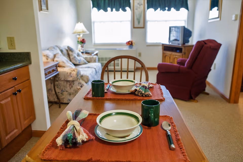 A cozy dining area with a wooden table set for two with green-rimmed bowls, plates, green mugs, and colorful napkins on orange placemats. In the background, there is a floral-patterned sofa, a wooden side table with a lamp, a burgundy recliner chair, and a small TV on a wooden cabinet. Two windows with green valances let in natural light.