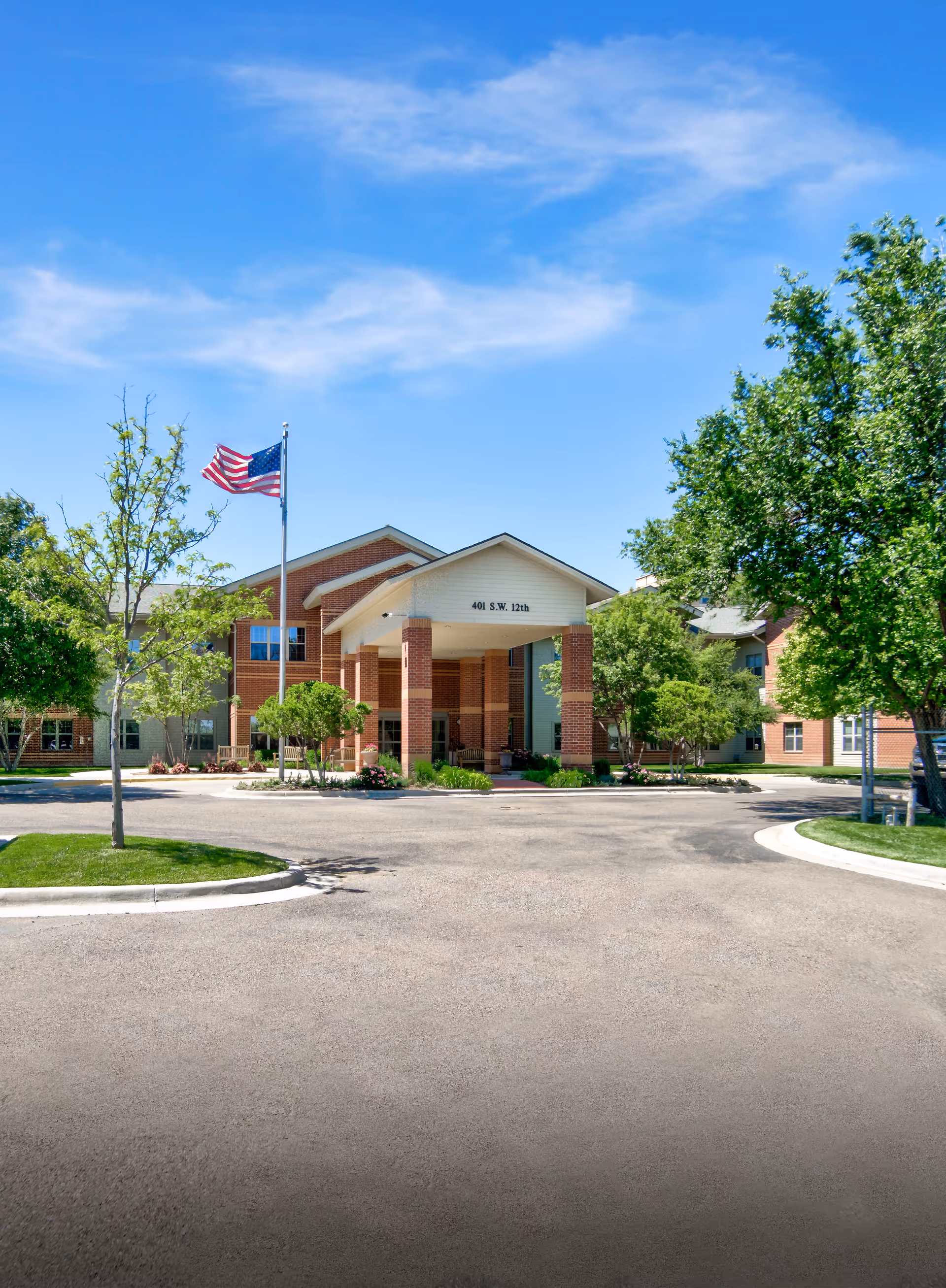 Front entrance of a red-brick senior living facility with a covered portico, American flag, and trees under a blue sky.