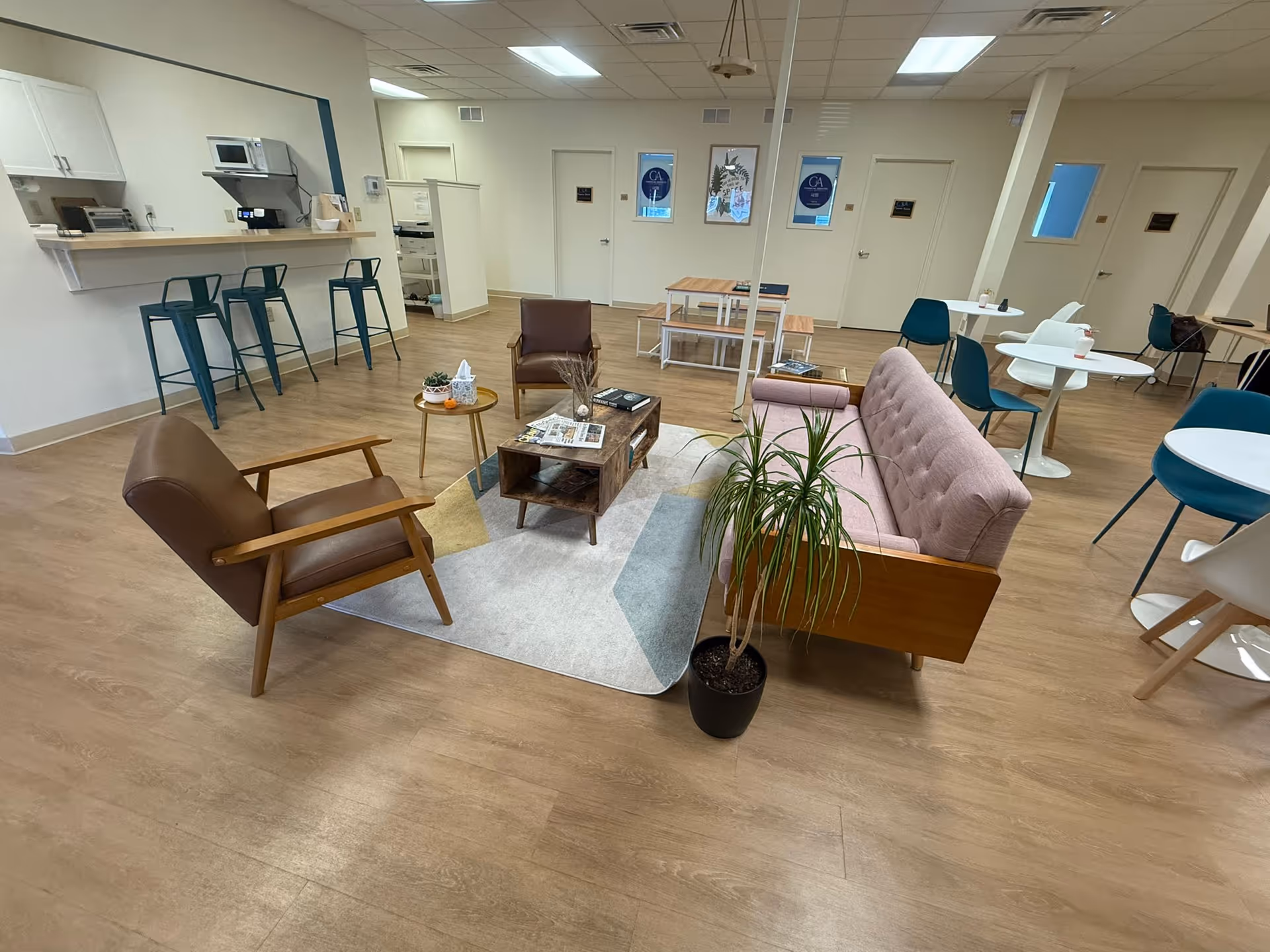 Modern senior living lounge with seating, tables, plant, and a kitchenette in the background.