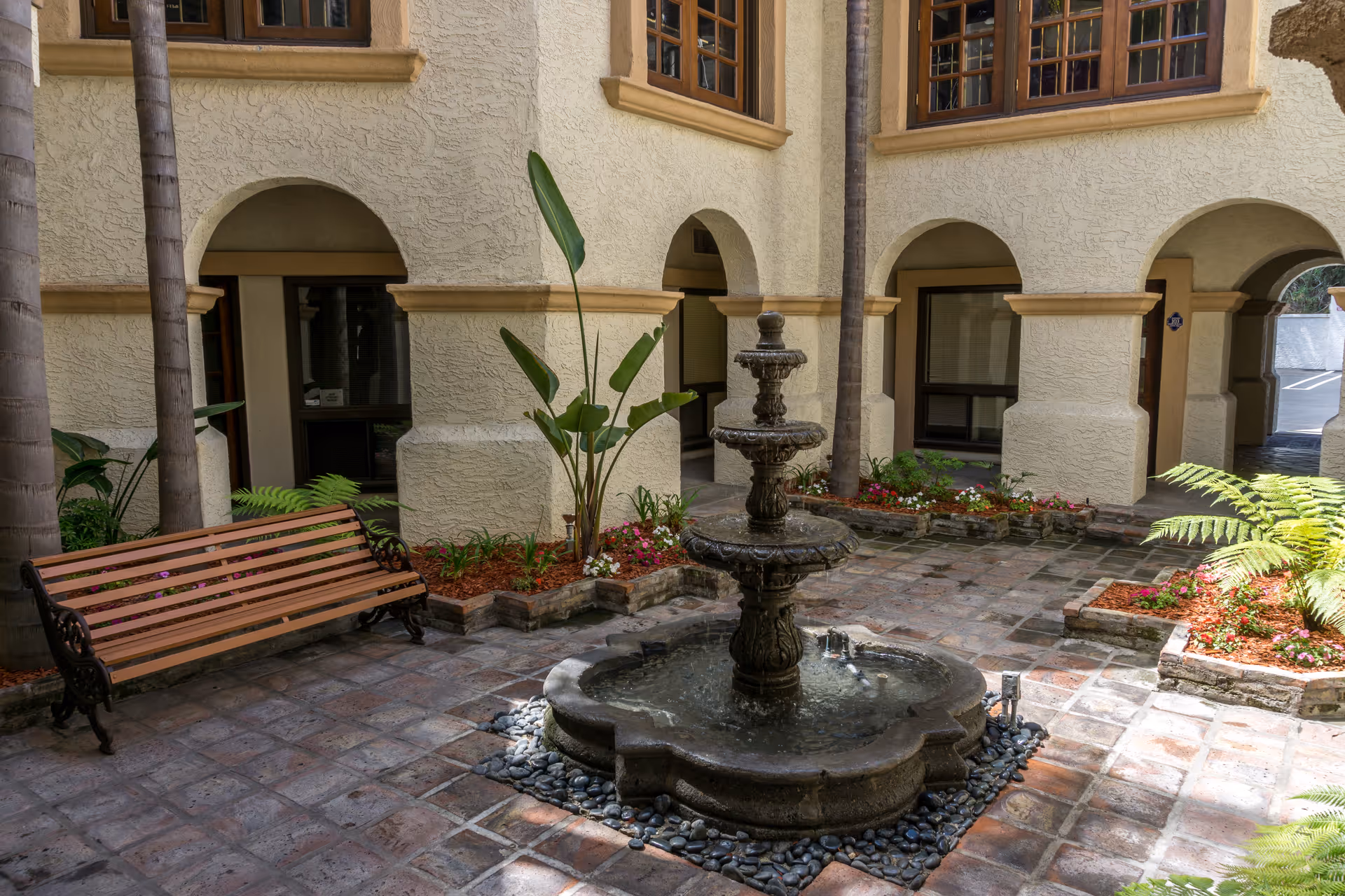 A stone courtyard with a central fountain, bench, arches, and tropical plants.