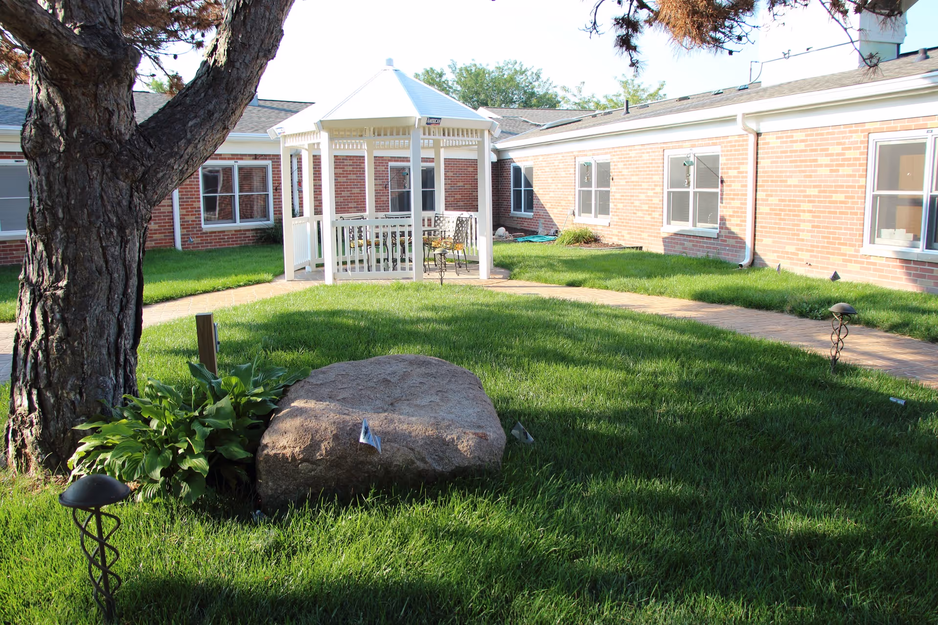 Brick senior living facility courtyard with a white gazebo, brick building, green lawn, and stone path.