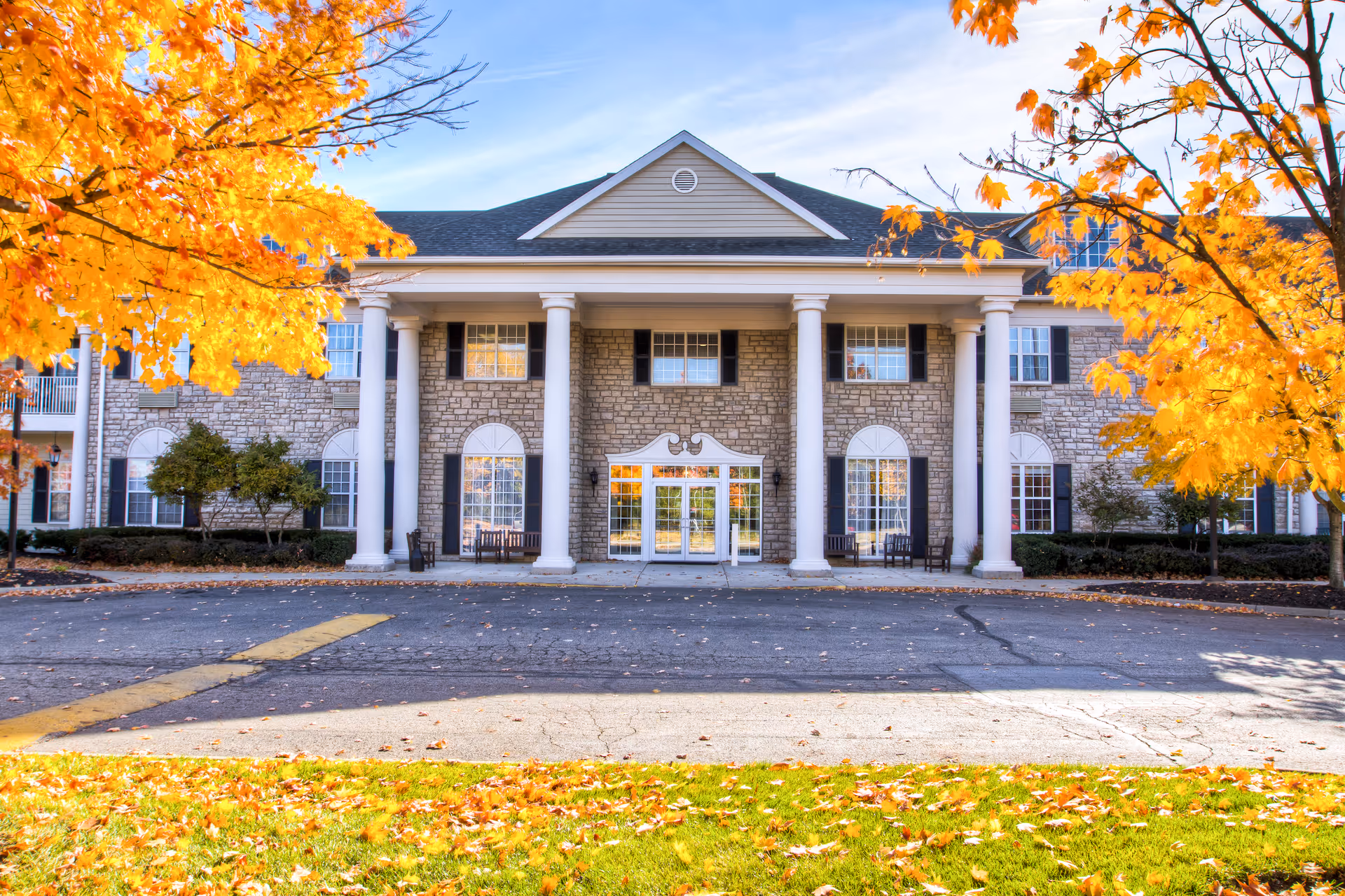Brookdale Muirfield exterior: stone facade, tall columns, glass doors, autumn trees framing entrance.