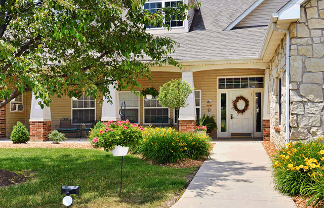 Front entrance of a senior living building with a walkway, porch, flowering shrubs, and a wreath on the door.