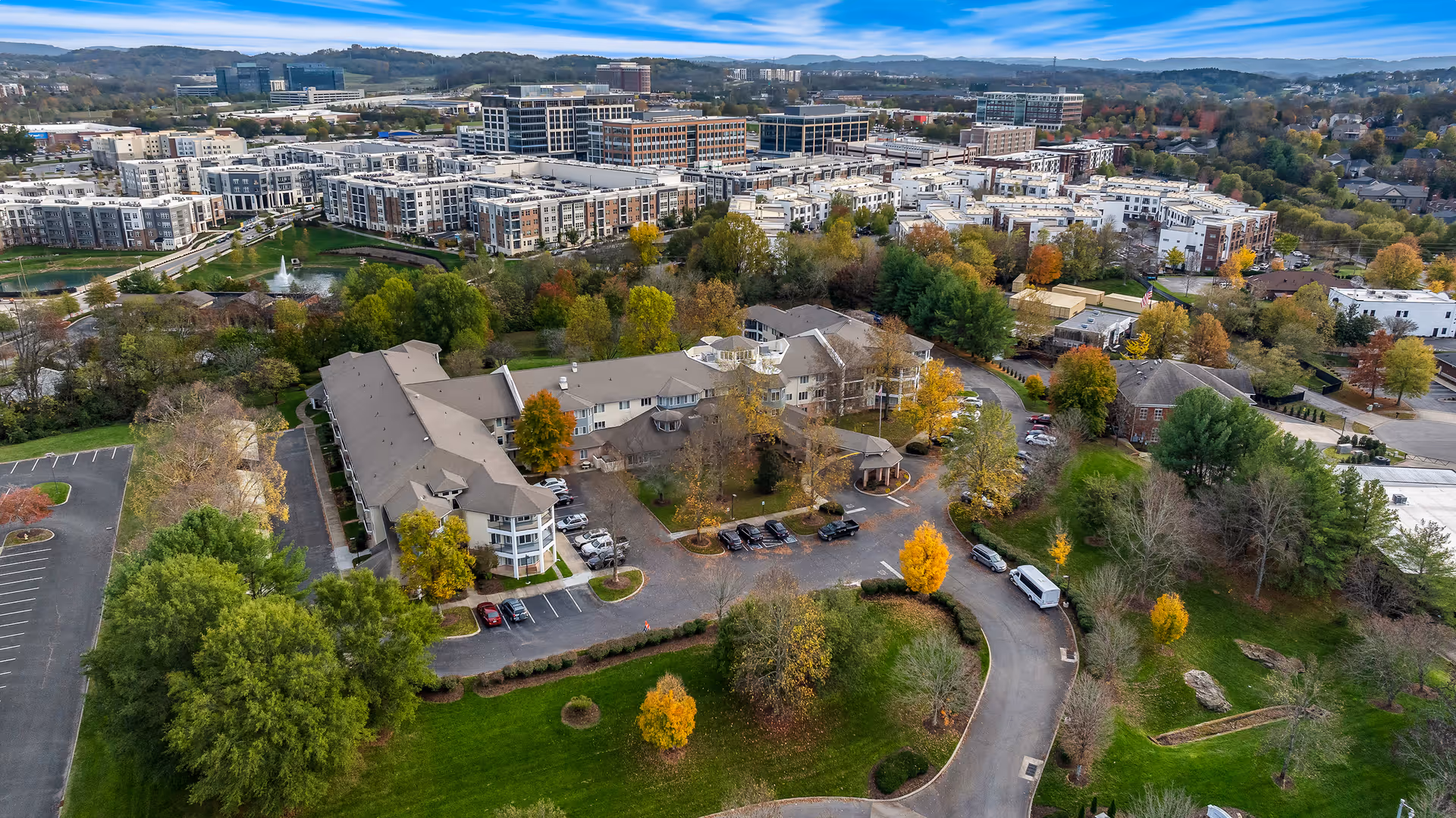 Aerial view of Manor at Steeplechase, a senior living community surrounded by trees and greenery, with a parking lot and a curved driveway. In the background, there is a large urban area with multiple buildings and a clear blue sky.