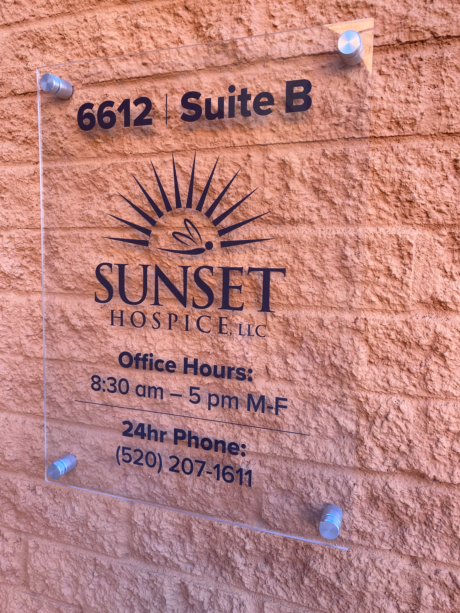 Exterior sign on orange stucco wall for Sunset Hospice LLC with hours and phone.