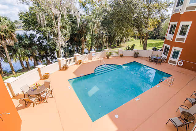 Outdoor swimming pool area at Brookdale Lake Orienta with a rectangular pool surrounded by a tan deck. There are tables with chairs and lounge chairs around the pool. The area is enclosed by a low wall, and there are trees and a body of water visible in the background.