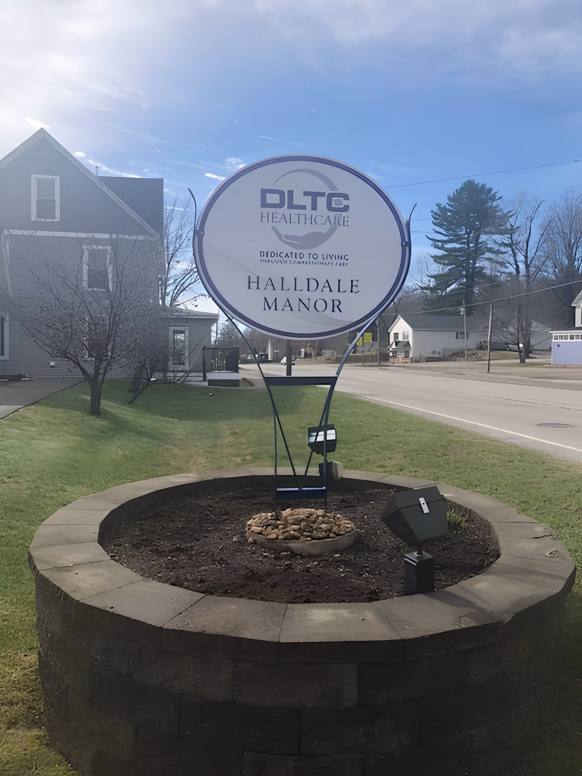 Circular DLTC Healthcare sign reading "Halldale Manor" above a round stone planter on the lawn in front of nearby houses and a street.