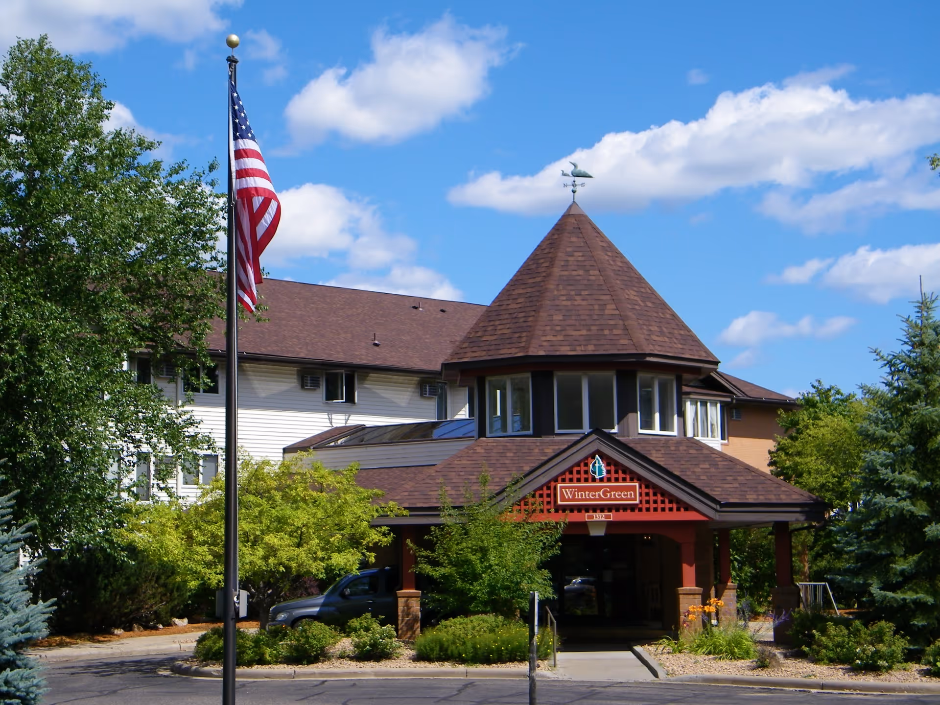 Exterior view of Wintergreen Independent Senior Living facility with a peaked roof entrance, surrounded by trees and shrubs, an American flag on a flagpole, and a partly cloudy blue sky.