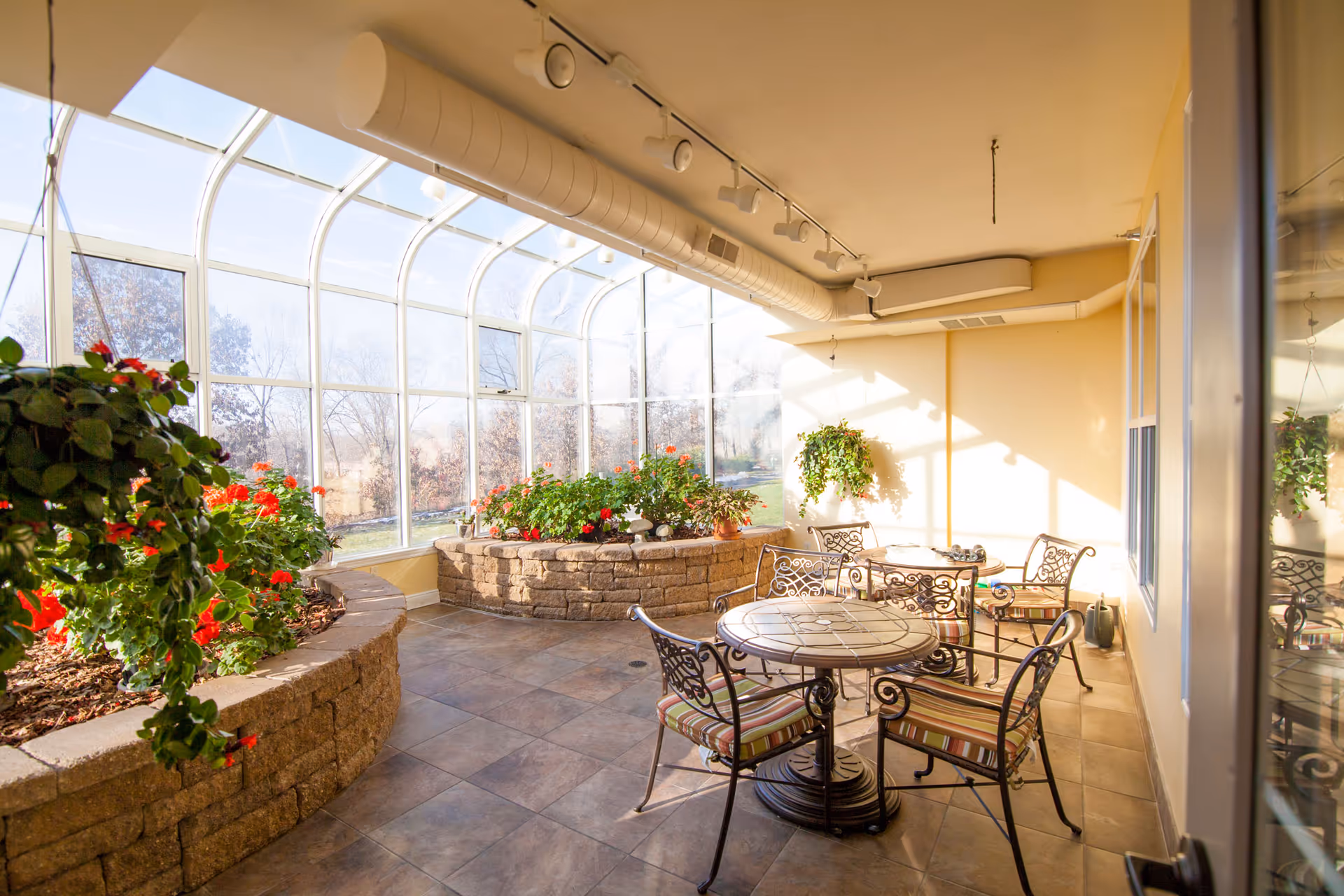 A bright indoor sunroom with large curved glass windows allowing natural light to fill the space. The room features two round tables with ornate metal chairs that have striped cushions. There are raised stone planters filled with green plants and red flowers, and a hanging plant on the wall. The floor is tiled, and the ceiling has exposed ductwork and track lighting.