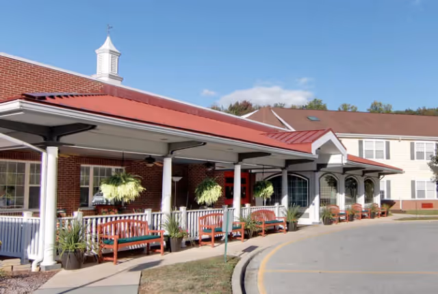Exterior view of Dover Place facility showing a covered entrance with a red metal roof, white columns, hanging plants, and wooden benches along the walkway. The building is made of red brick and light-colored siding under a clear blue sky.