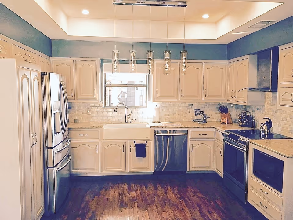 A modern kitchen with cream-colored cabinets, stainless steel refrigerator, dishwasher, oven, and microwave. The kitchen features a farmhouse sink under a window, pendant lights hanging from the ceiling, and wooden flooring.