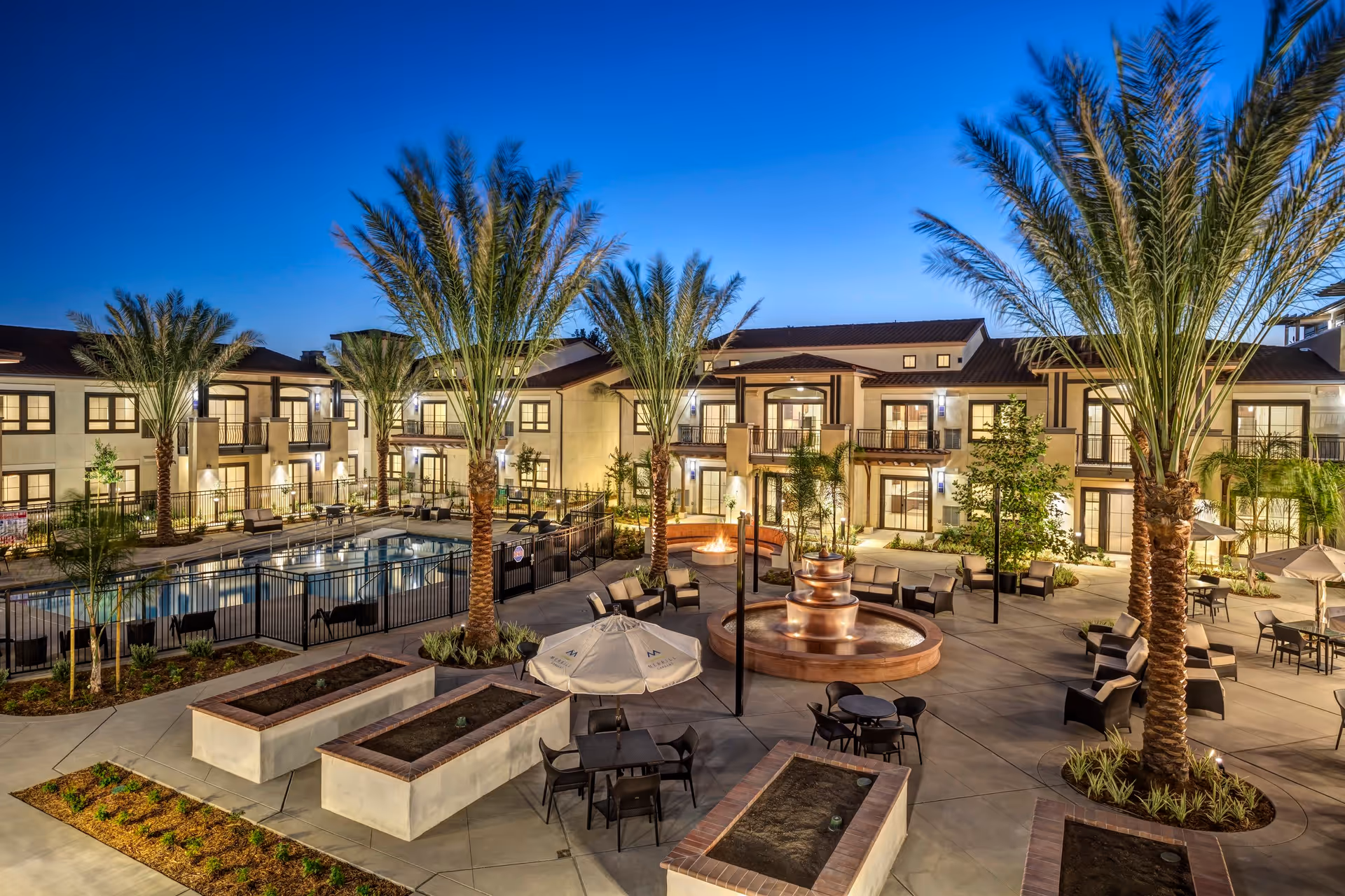 Evening view of a landscaped courtyard with palm trees, a central fountain, pool, seating areas and surrounding apartment-style building.