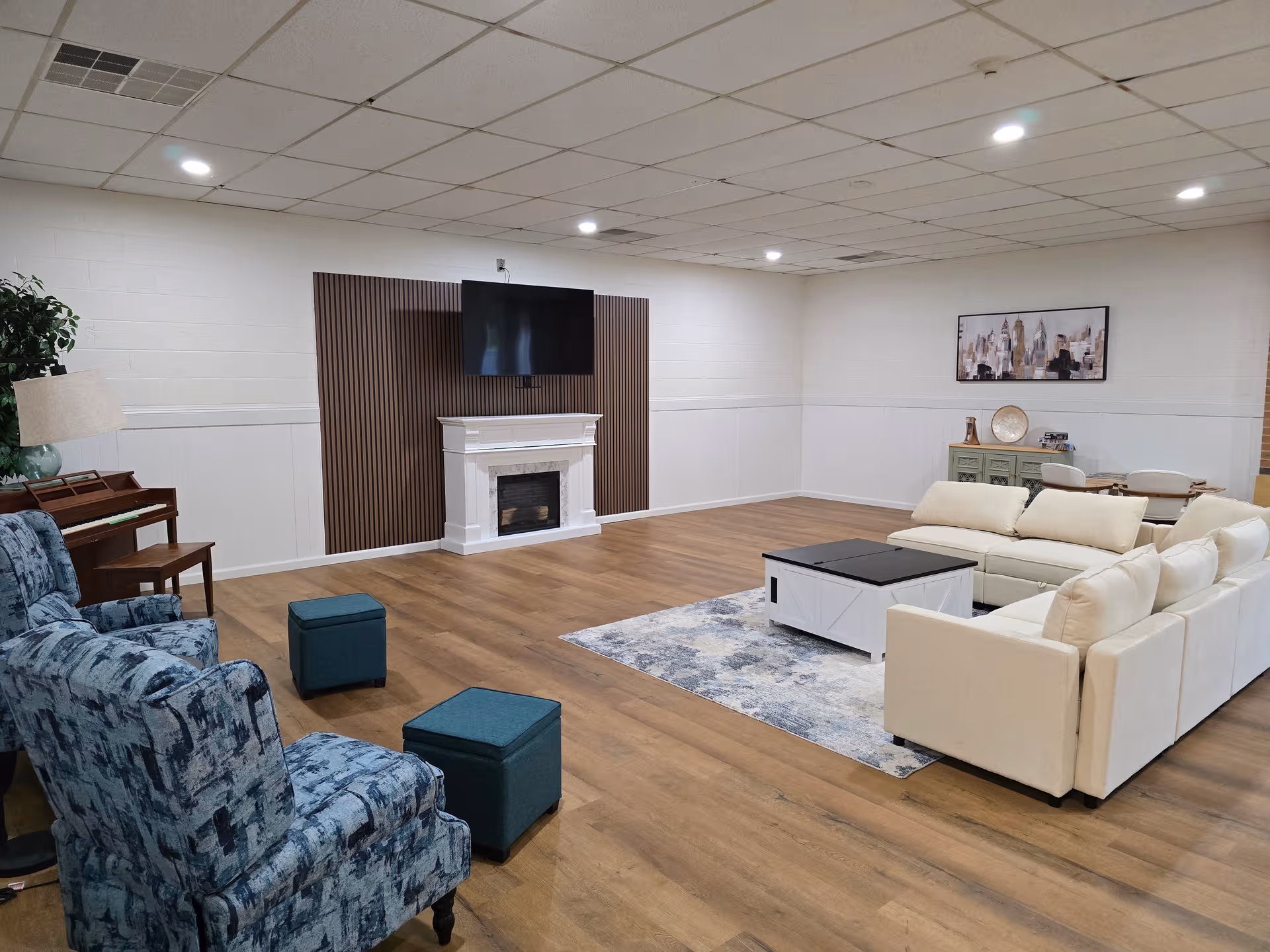 Spacious senior living lounge with white sectional sofa, fireplace, piano, and mounted TV.