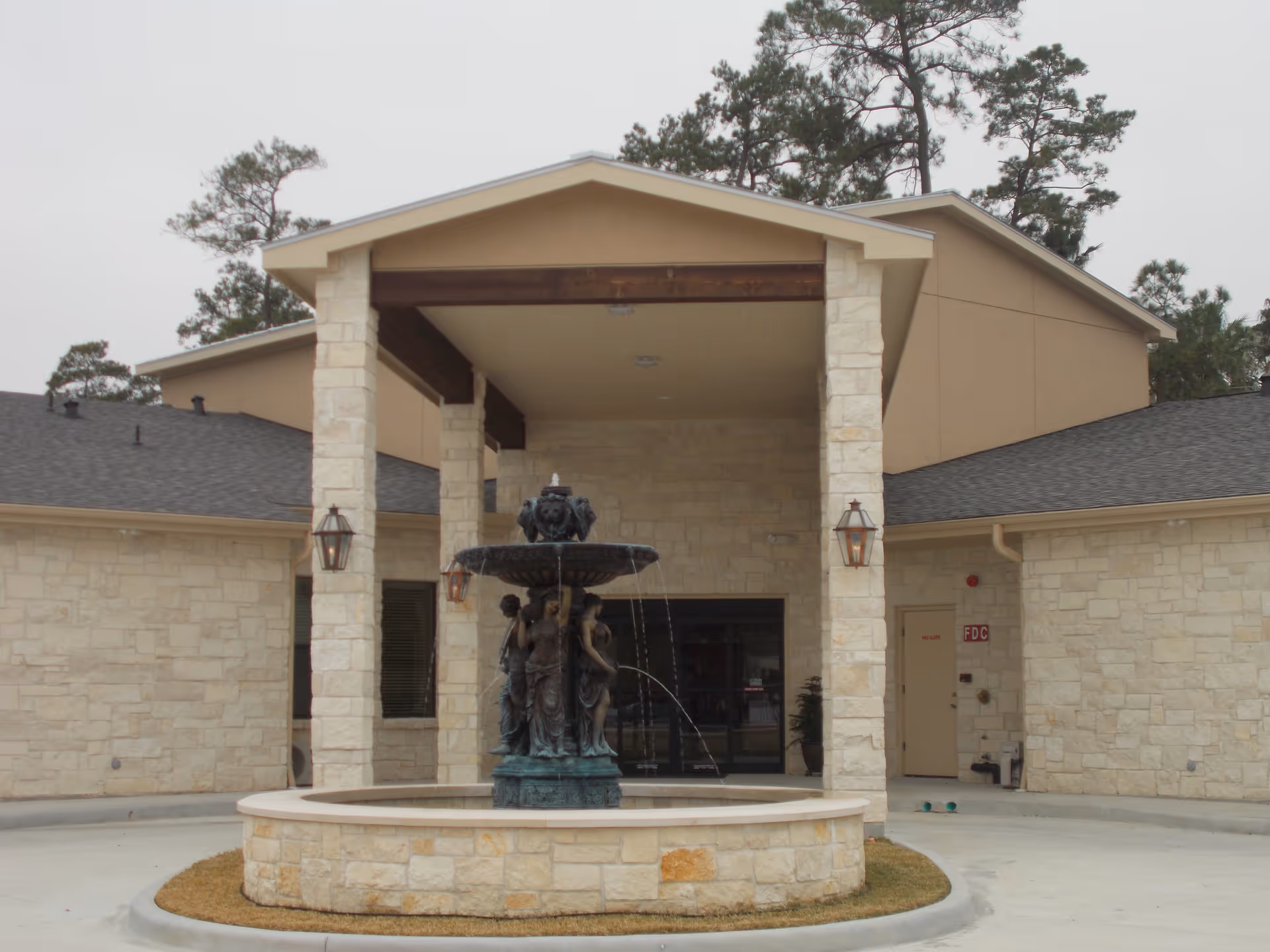 Entrance of a senior living facility with a covered porch supported by stone pillars and a decorative water fountain in the circular driveway in front. The building exterior is made of light-colored stone with a beige upper section and a dark shingled roof. Trees are visible in the background.