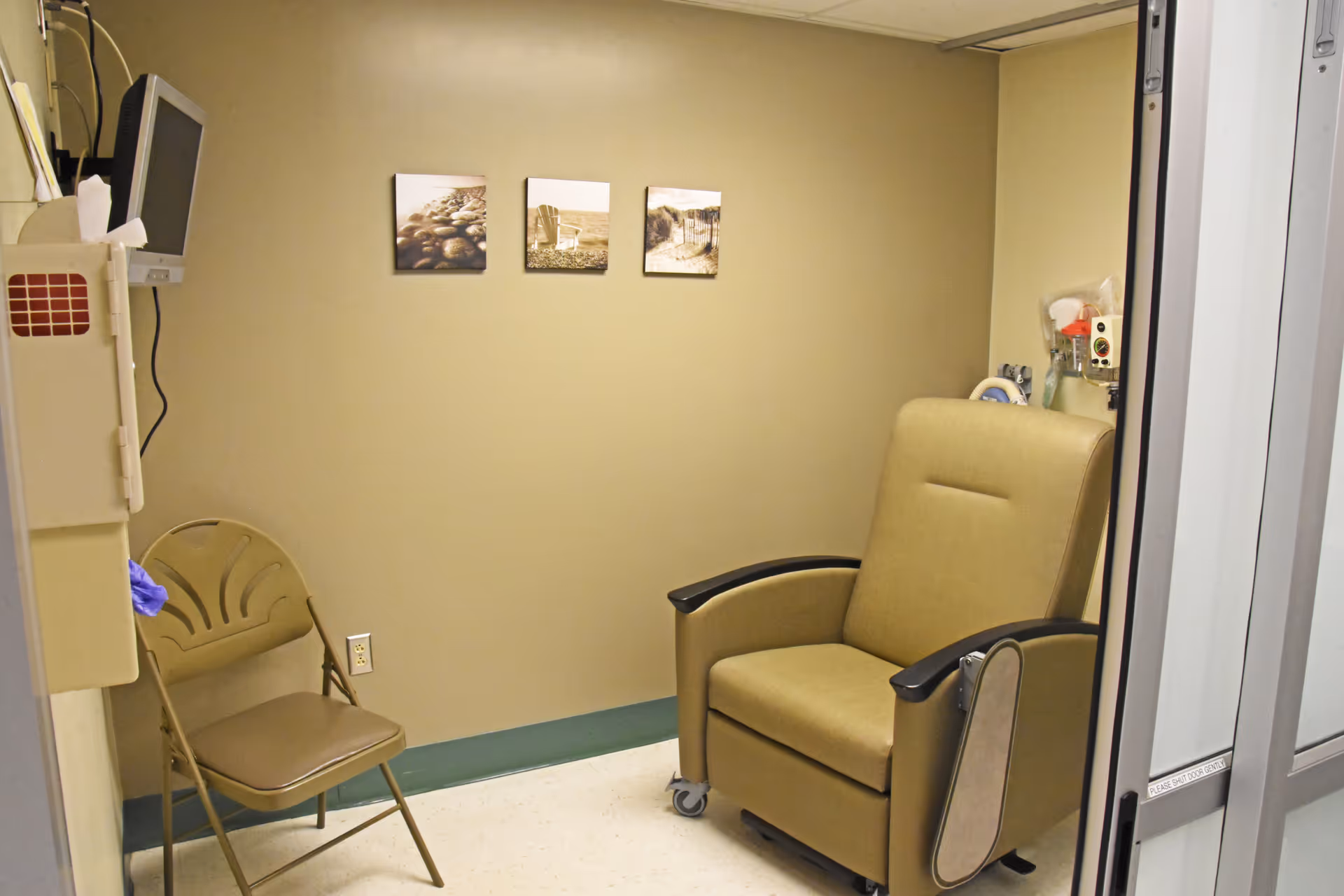 A beige medical recliner chair sits beside a folding chair in a small treatment room.