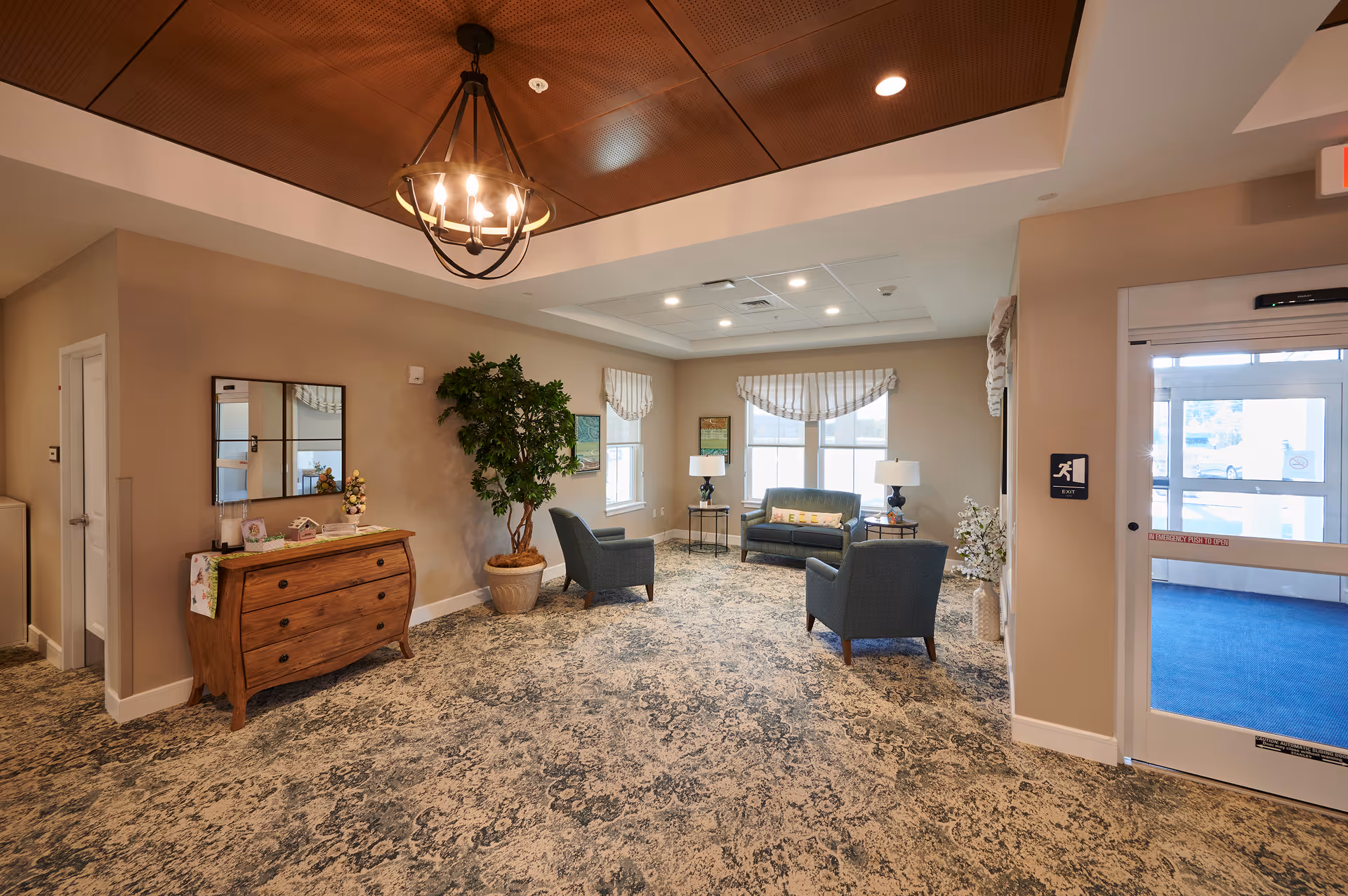 A cozy and well-lit sitting area in a senior living facility with a patterned carpet, two armchairs, a small loveseat, side tables with lamps, a large potted plant, a wooden chest of drawers with decorative items, and a chandelier hanging from a wooden ceiling panel. There is a glass door leading outside and windows with valances letting in natural light.