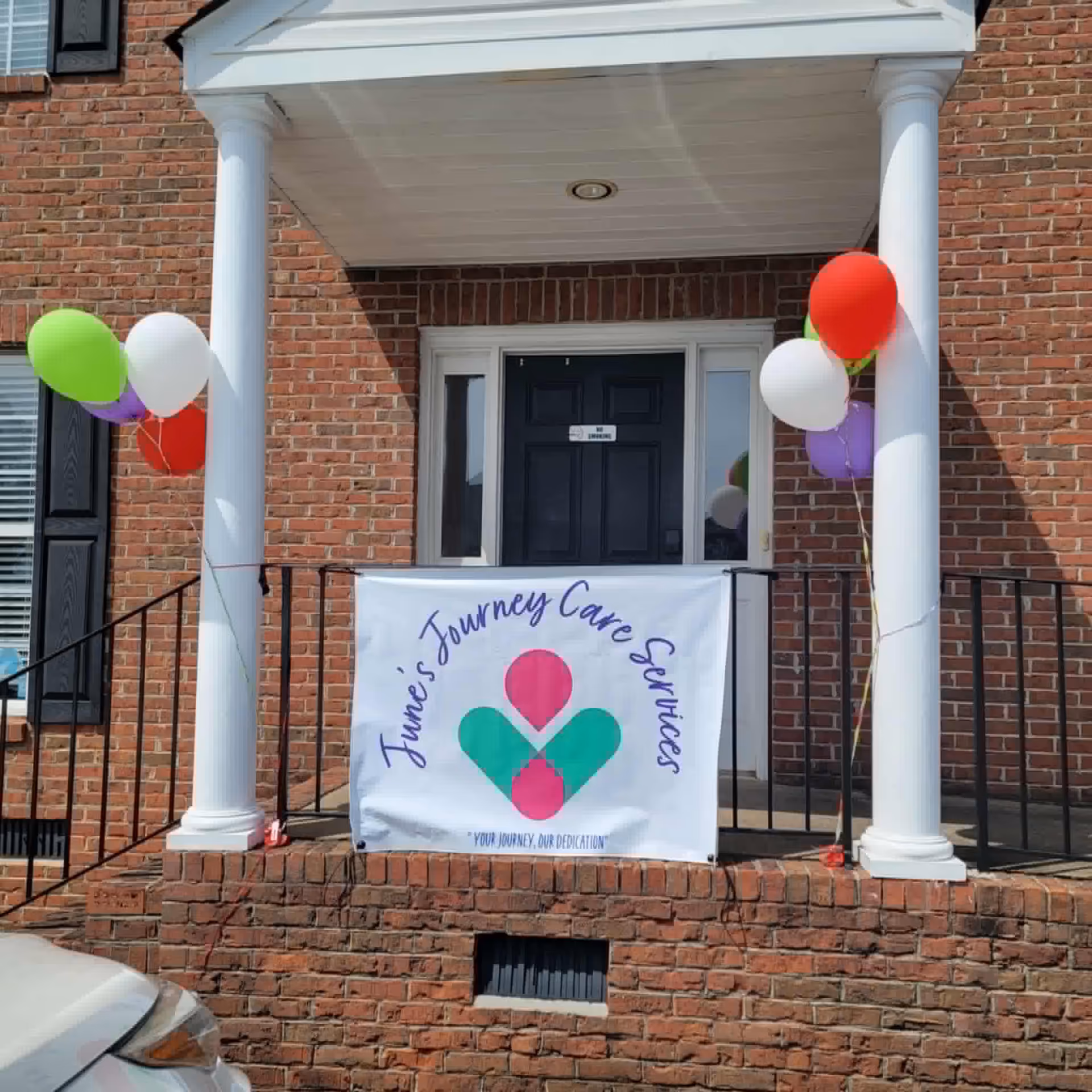 Brick building entrance with white columns, balloons, and a banner reading June's Journey Care Services.