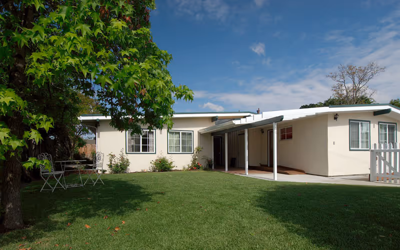 Front view of a single-story cream-colored building with a covered entrance, green lawn, and a tree with a small outdoor table and chairs.