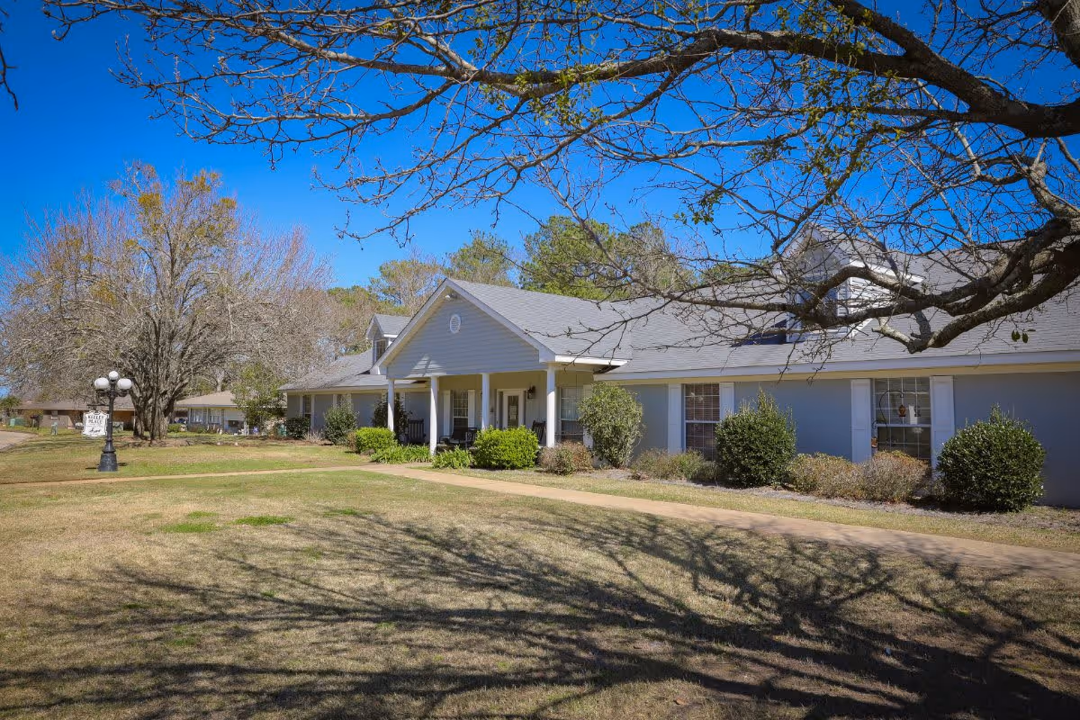 Exterior view of Kelley Place, a single-story building with a gray roof and light blue walls, surrounded by bushes and trees under a clear blue sky. A paved walkway leads to the entrance with white columns and a small porch. A lamp post with a sign reading 'Kelley Place' is visible on the left side of the image.
