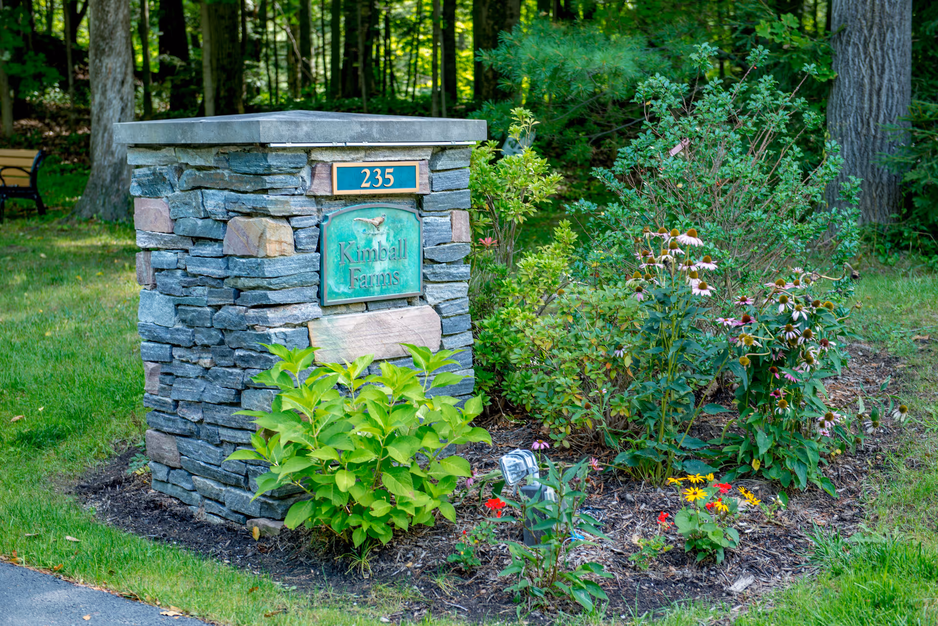 A stone pillar with a sign reading 'Kimball Farms' and the number 235, surrounded by green plants and flowers, with a wooded area in the background.