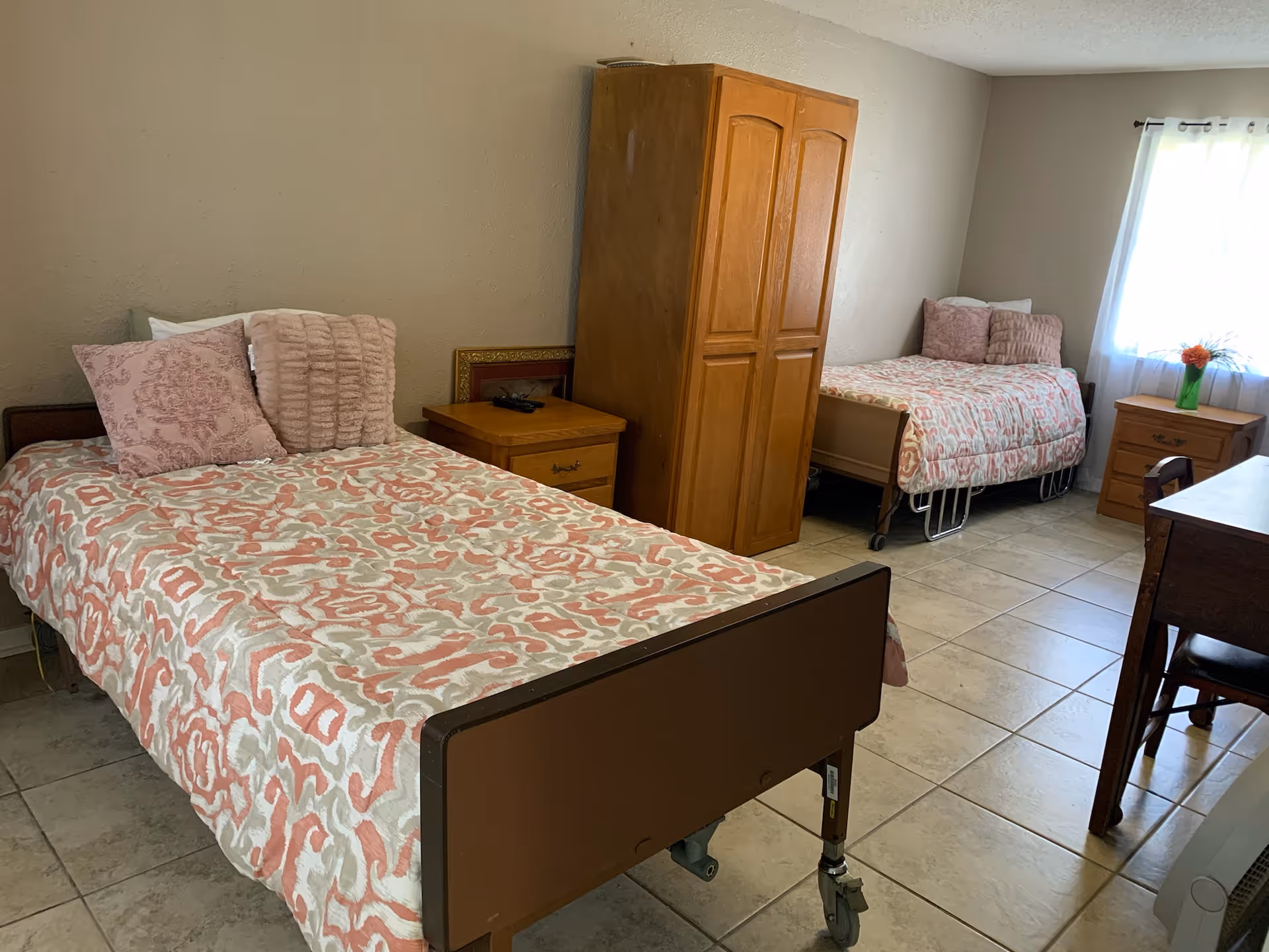 Two twin beds with pink bedding, a wardrobe, and nightstands in a shared senior living bedroom.