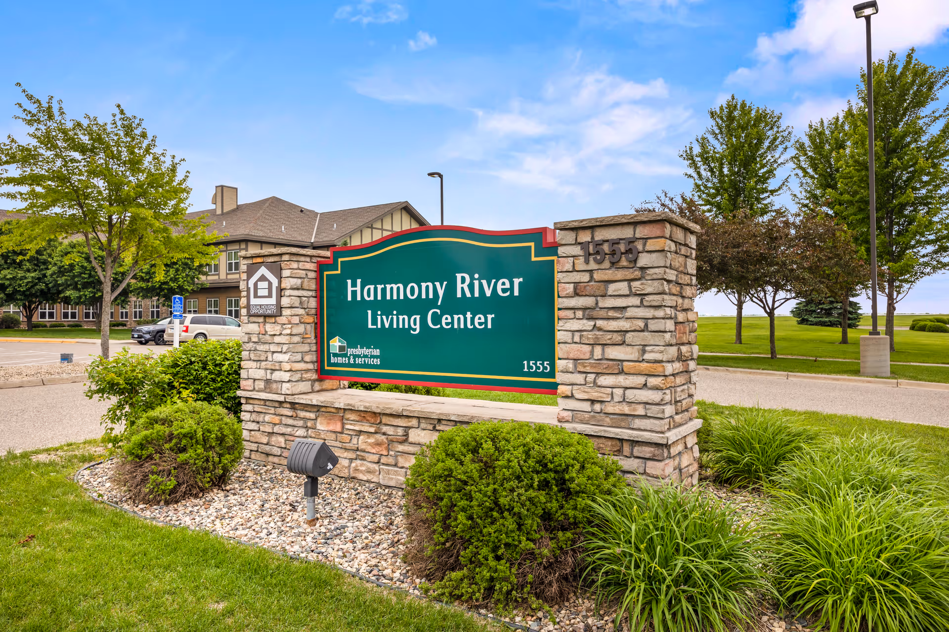 Stone sign for Harmony River Living Center with landscaping and a building in the background under a partly cloudy sky.