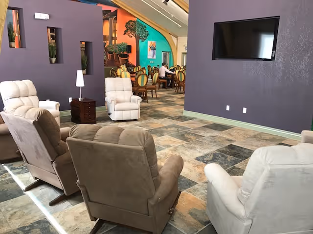 A spacious lounge area with several cushioned recliner chairs arranged facing a wall-mounted flat screen TV. The floor is tiled with a mix of earth tones, and the walls are painted purple. In the background, there is a dining area with tables and chairs, and a colorful mural on the far wall.