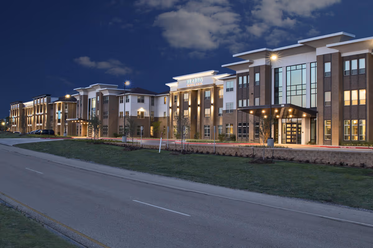 Illuminated three-story Heartis Mid Cities senior living building at night with a covered entrance and landscaped front lawn.