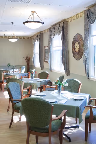 A bright dining room with several tables covered in green tablecloths, each set with white plates, napkins, and silverware. Green upholstered chairs with wooden arms surround the tables. The room has large windows with patterned curtains, a decorative wall clock, and ceiling lights providing illumination.