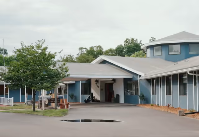 Blue, single-story assisted living facility exterior with entrance canopy and surrounding trees.