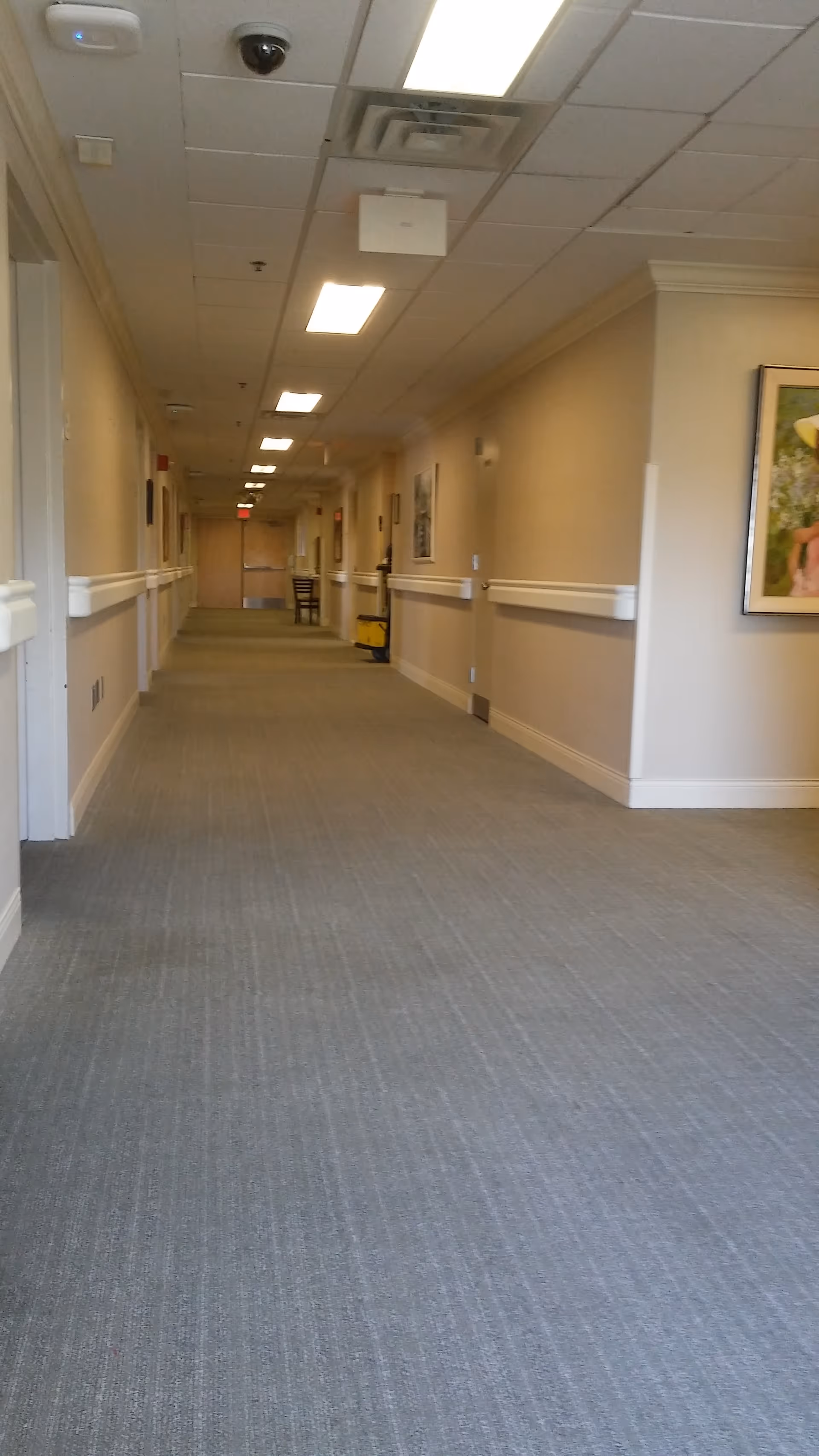 Long interior hallway in a hospice facility with beige walls, ceiling lights, and railings.