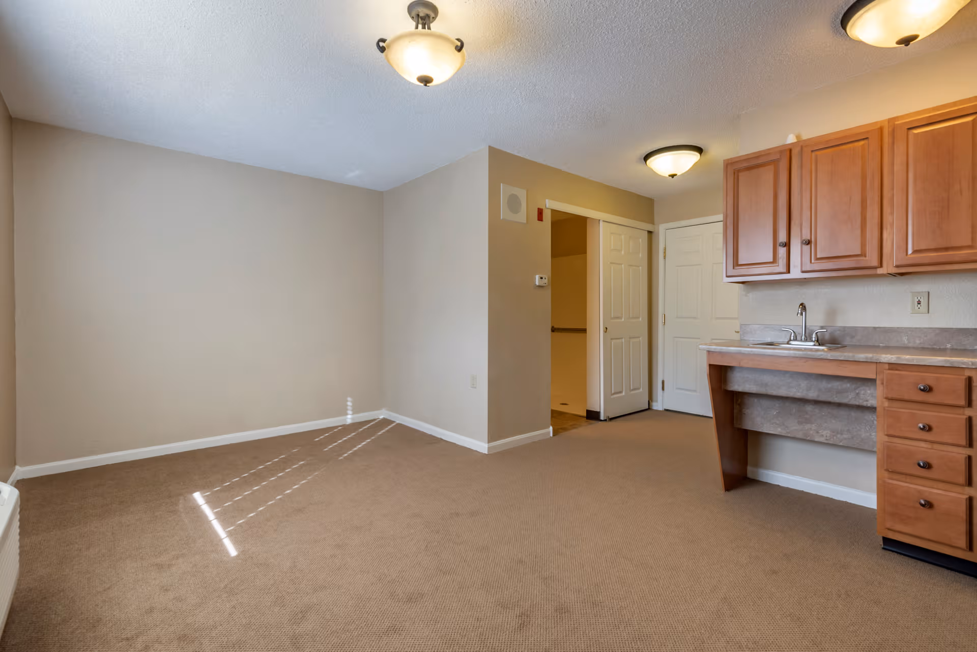 Neutral-toned kitchen and living area with wooden cabinets, sink, and carpeted floor.