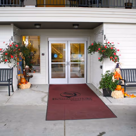 Entrance of a senior living building with double glass doors, a maroon welcome mat branded 'Bonaventure,' benches and fall decorations.