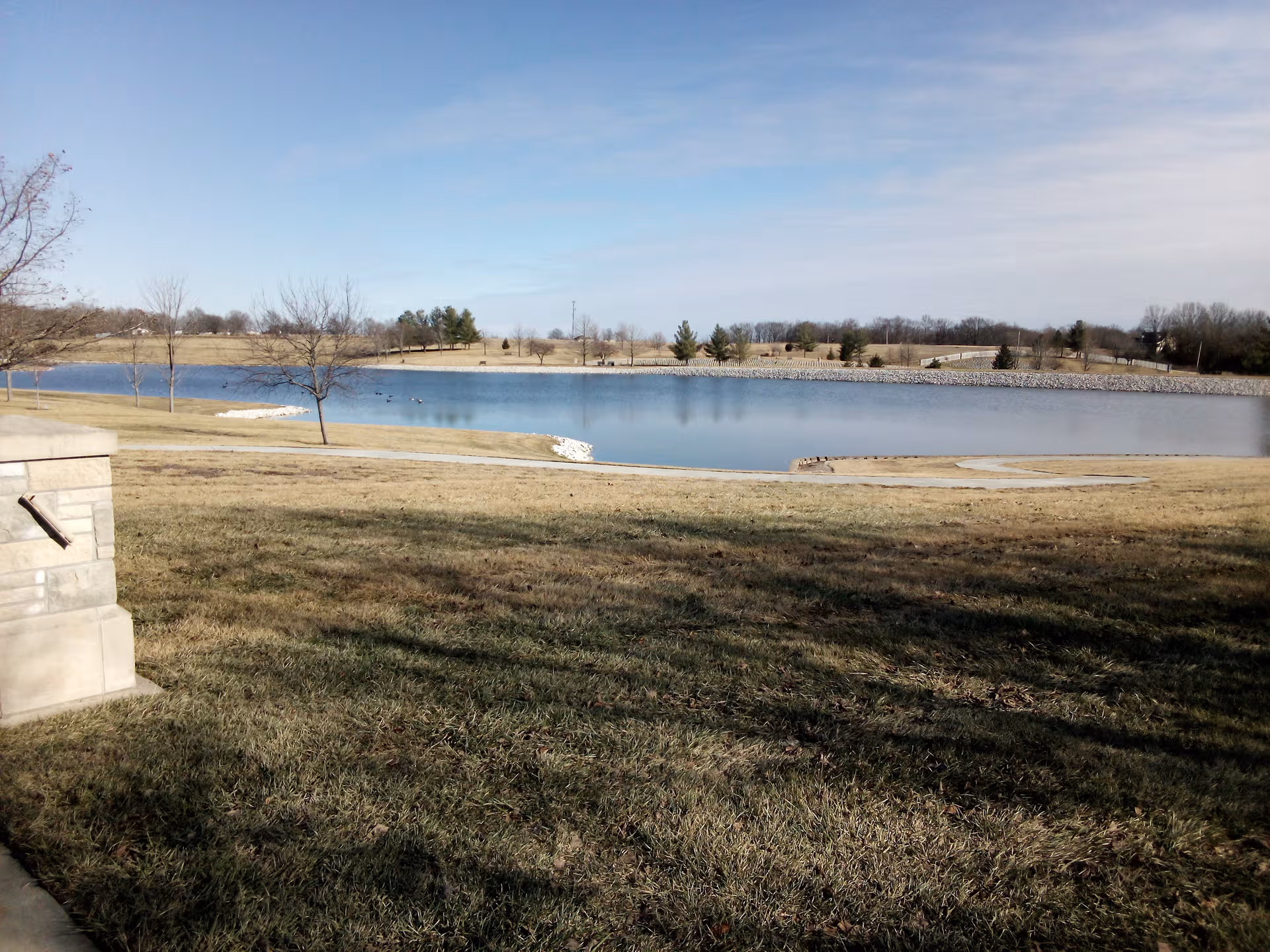 Expansive grassy park with a calm lake, bare trees, and a clear sky.