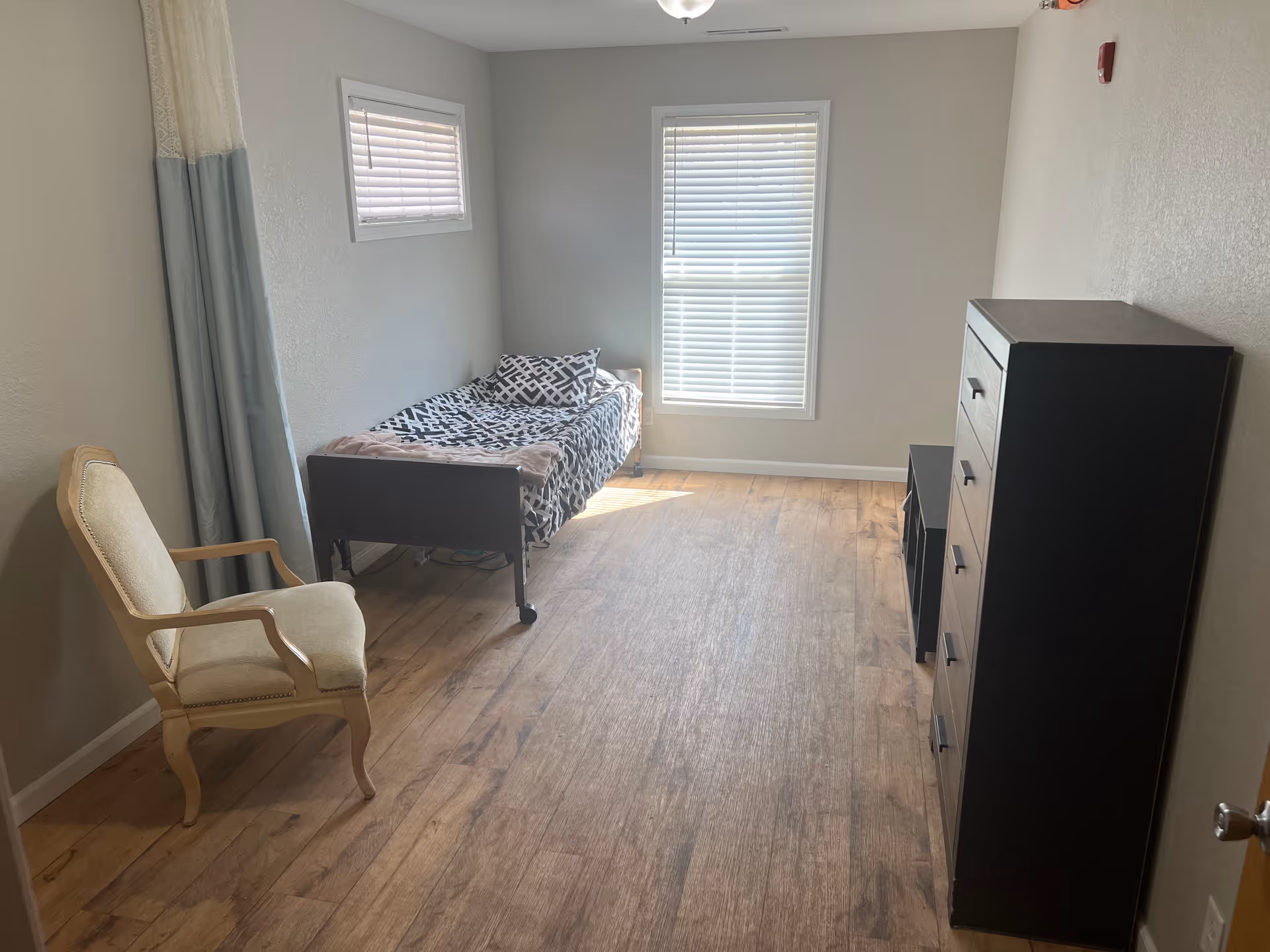 A sparsely furnished bedroom in a senior living facility with a bed, chair, and dresser.