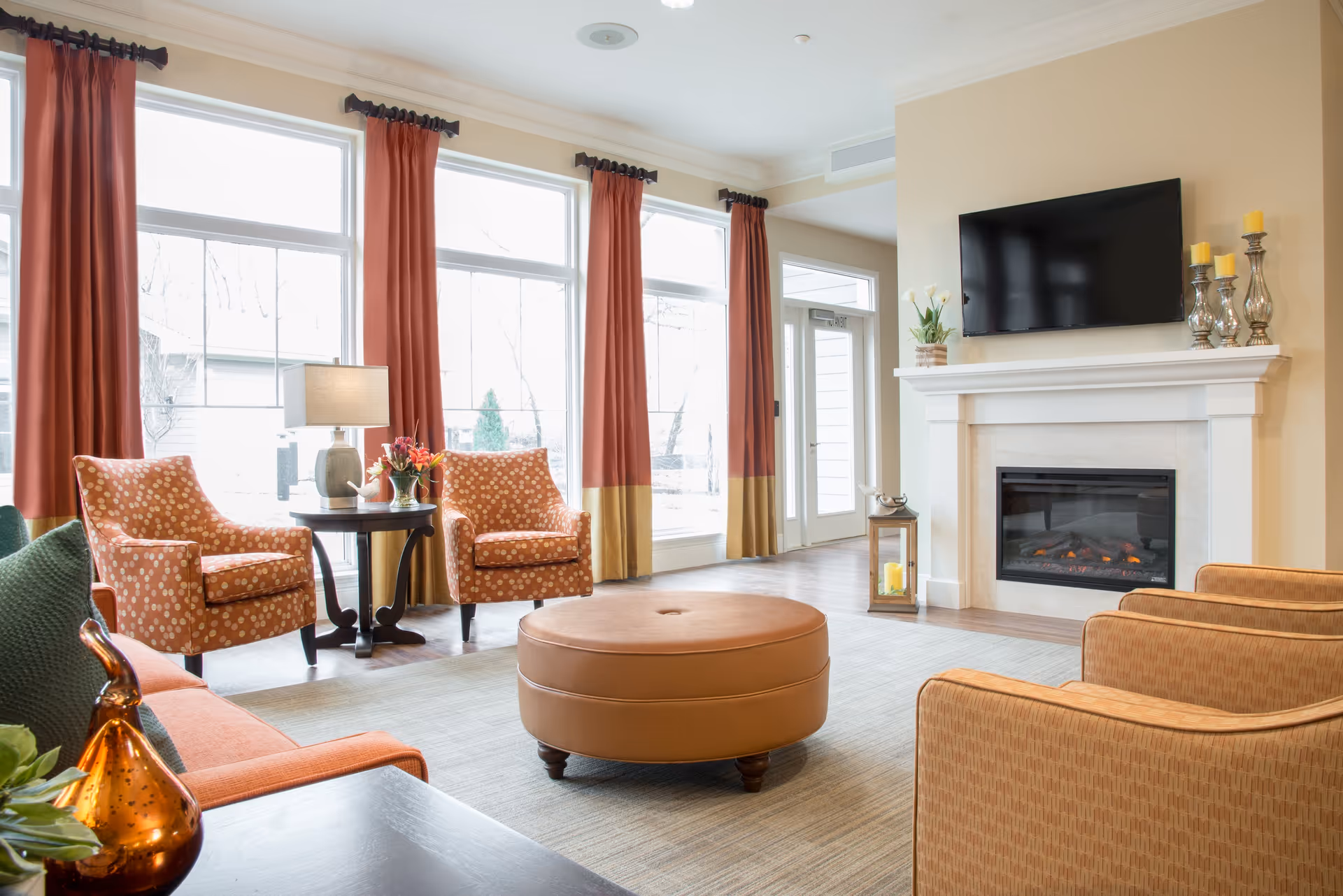 A bright and cozy living room with large windows covered by red and gold curtains, two patterned armchairs around a small round table with a lamp and flowers, a large round leather ottoman in the center, a fireplace with candles and a flat-screen TV mounted above it, and additional seating with orange upholstered chairs.