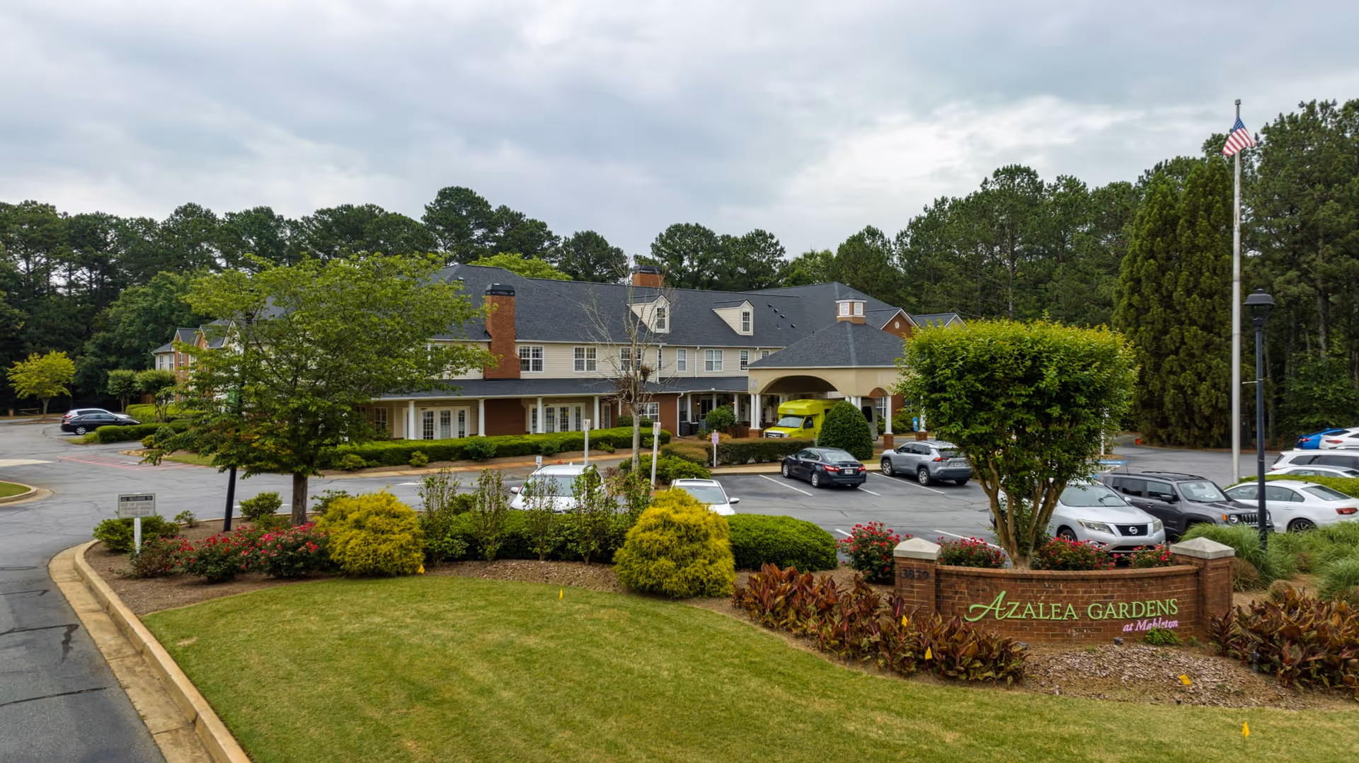 Exterior view of Azalea Gardens Mableton senior living facility showing a large two-story building with a covered entrance, surrounded by trees, shrubs, and a parking lot with several cars. A brick sign with green and pink lettering reading 'Azalea Gardens at Mableton' is visible in the landscaped front area.