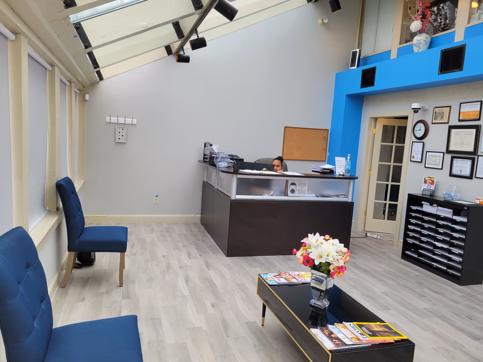 Reception desk with a staff member behind, blue chairs, flowers, and certificates in a well-lit lobby