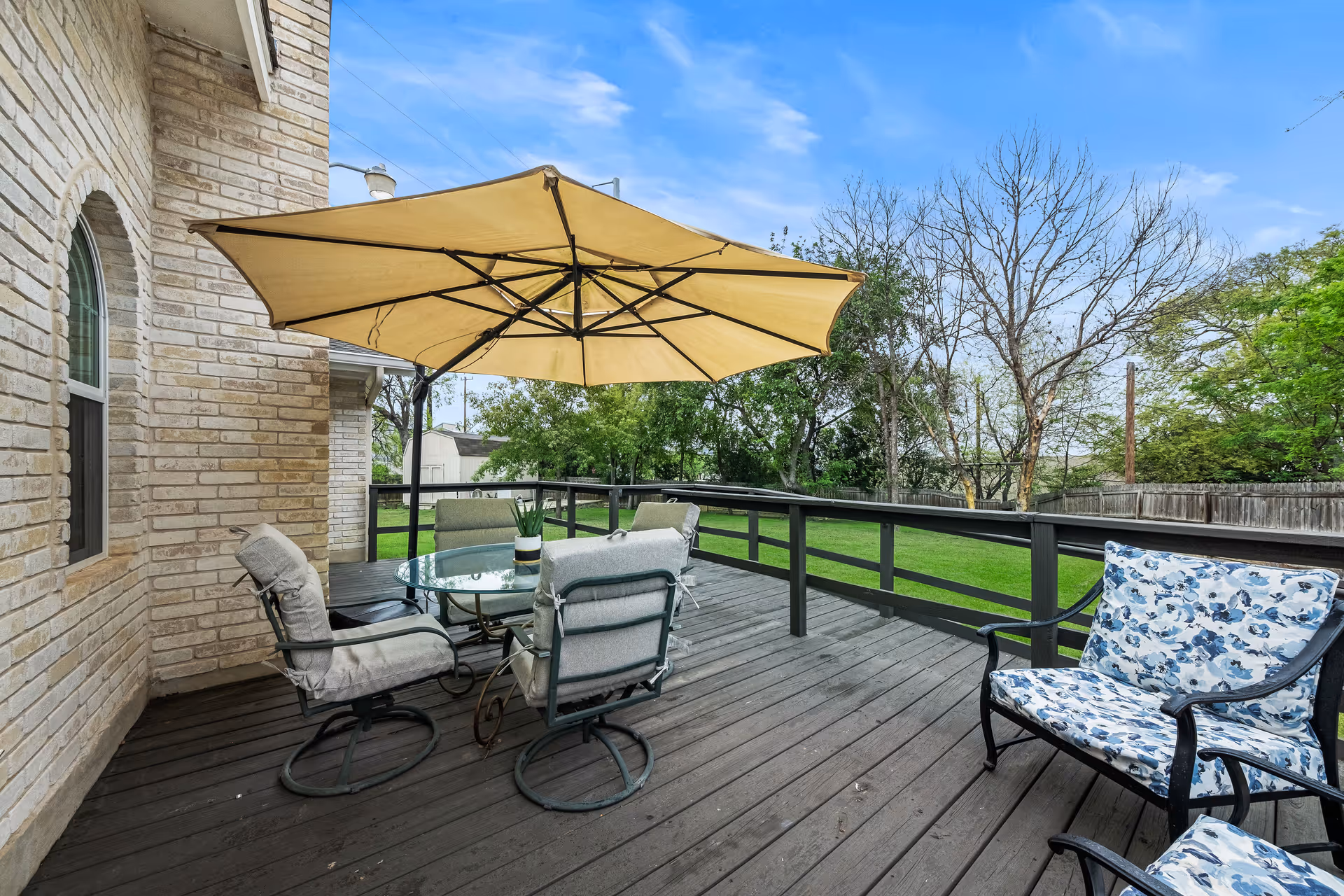 Outdoor wooden deck attached to a brick building with a round glass table surrounded by four cushioned chairs and a large beige patio umbrella. There is also a cushioned bench with a blue and white floral pattern. The deck overlooks a green lawn with trees and a wooden fence under a blue sky with some clouds.