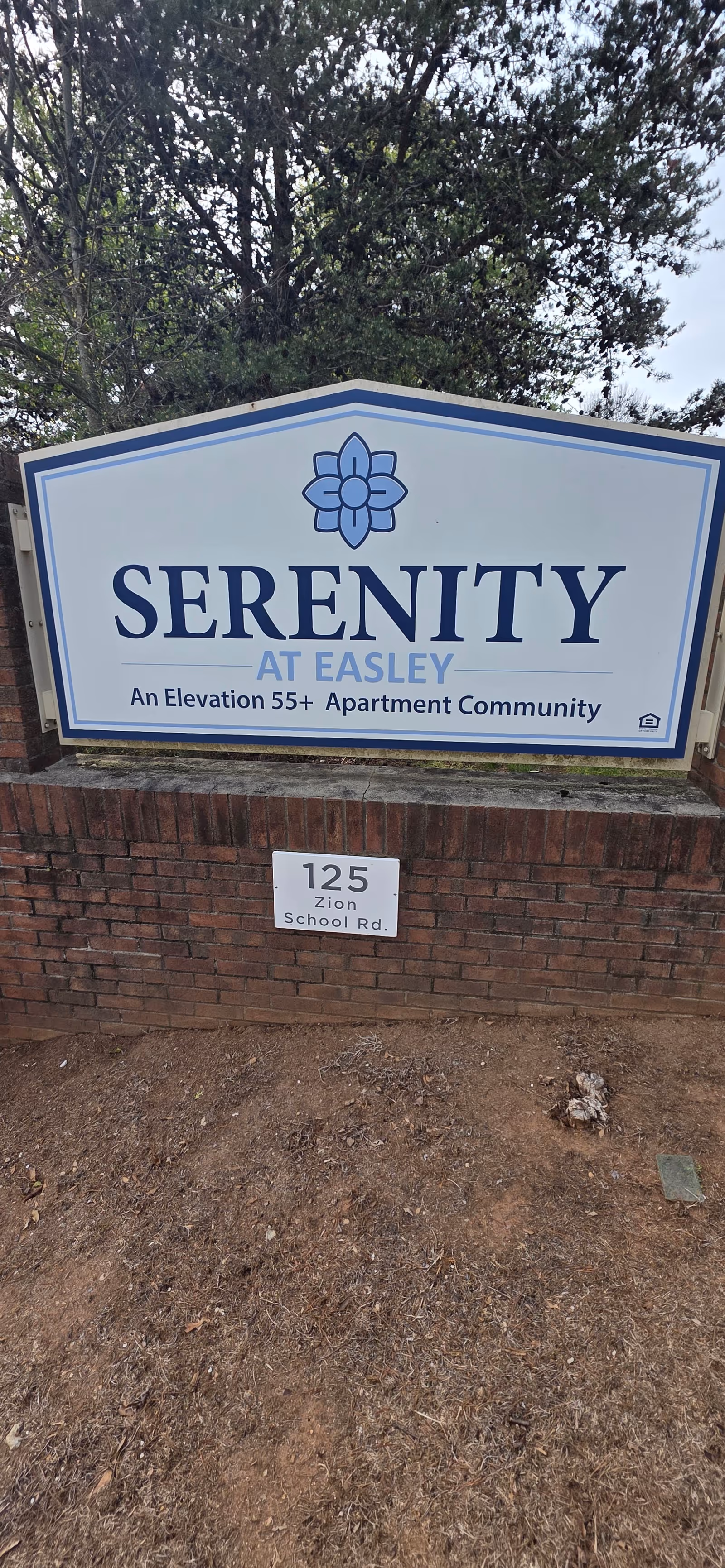 Sign reads Serenity at Easley, An Elevation 55+ Apartment Community, brick base with trees behind.