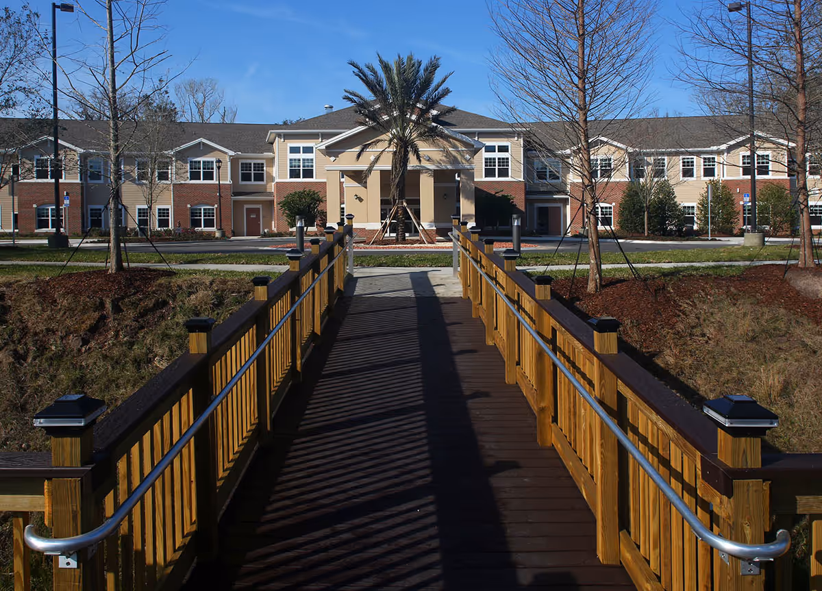 A wooden pedestrian bridge with handrails leading to the entrance of a two-story senior living facility building with beige and brick exterior walls, surrounded by trees and landscaping under a clear blue sky.